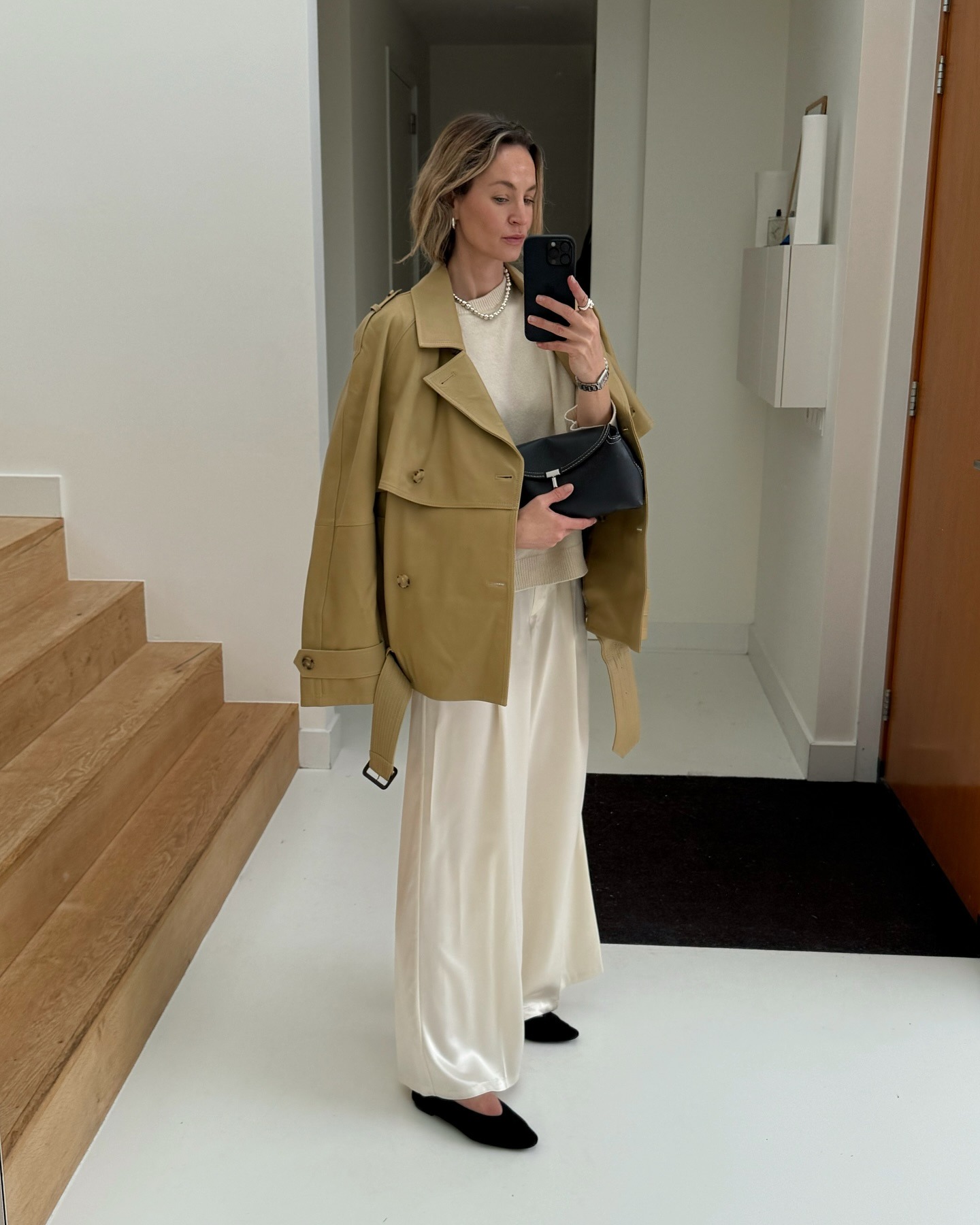 Dutch style influencer Anouk Yve poses for a mirror selfie wearing a cropped trench coat, beige sweater, black Toteme clutch, white satin pants and black ballet flats