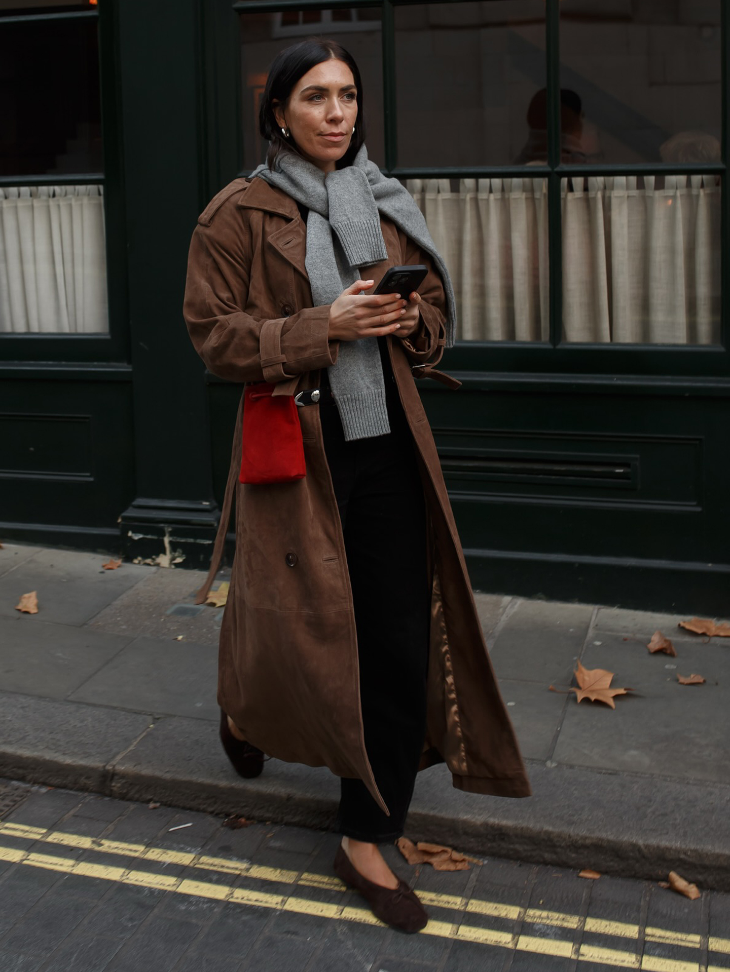 British influencer Jessica Skye walking. inLondon wearing a long brown suede trench coat, a gray sweater tied around the neck, a red suede bag, black barrel jeans, and brown v-cut flats