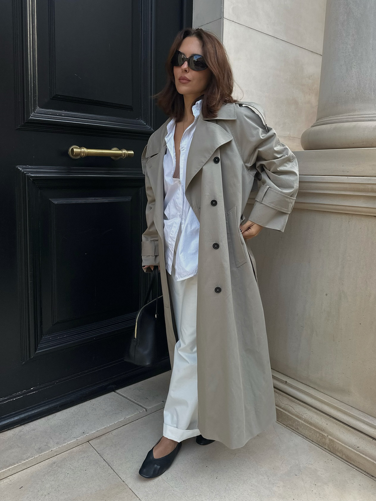 Style influencer Debora Rosa poses in a long gray trench coat, white button-down shirt, ivory cuffed pants, a black bag, and black v-cut flats