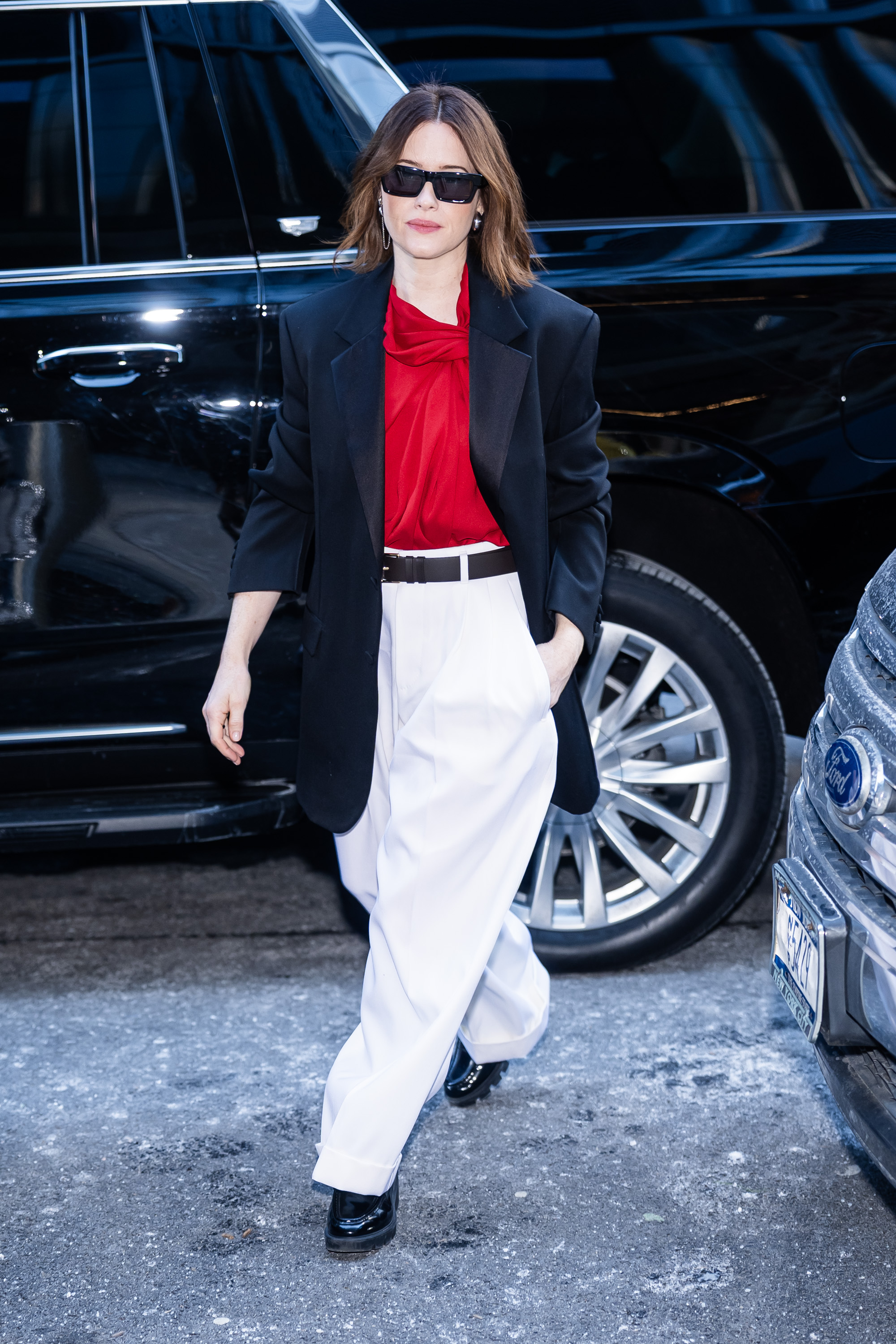 Claire Foy steps outside a car wearing white trousers with black patent flats, a red blouse and a black blazer.