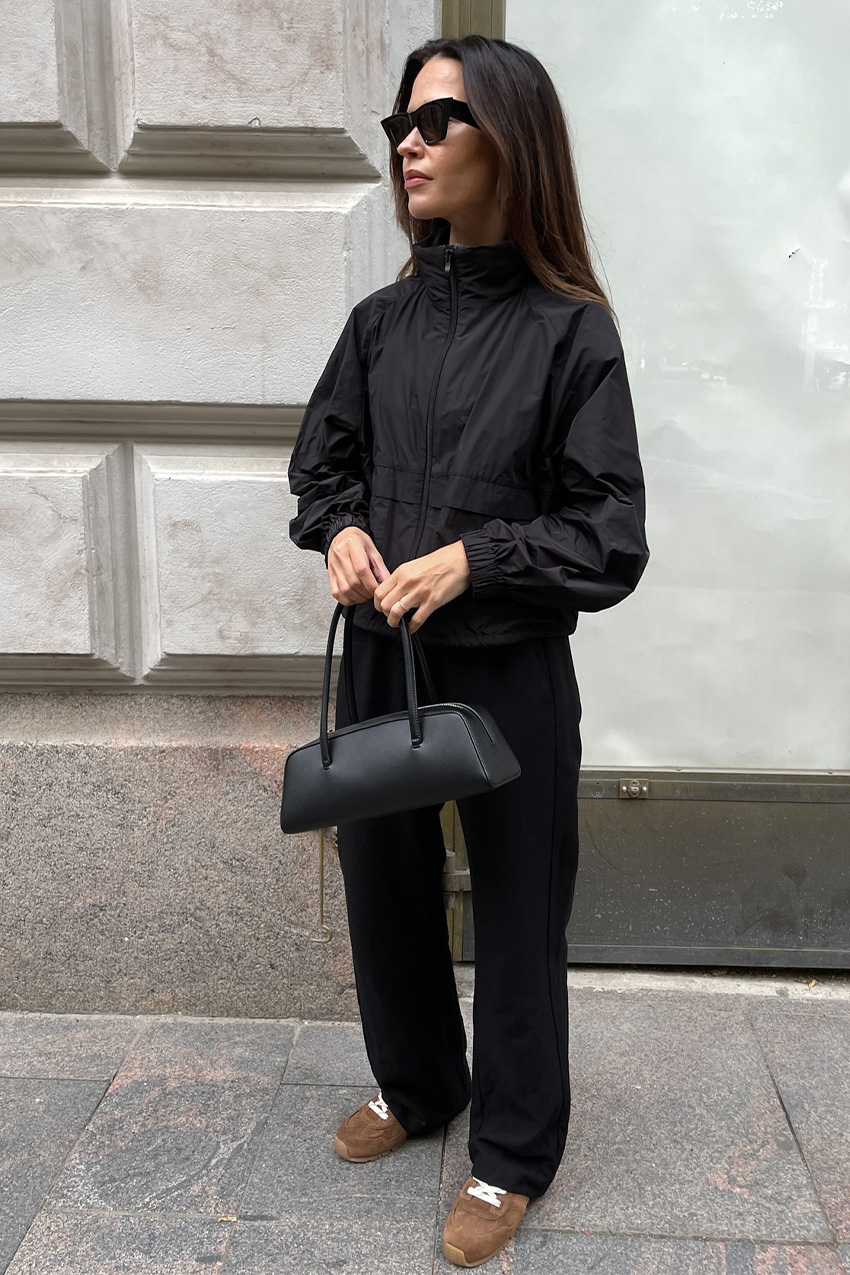 Shoe trends to wear with leggings, jeans, and skirts in 2026 are shown in a street style photo of a woman standing on the sidewalk in front of a concrete wall wearing black sunglasses, a black funnel neck rain jacket, black trousers, a black baguette, and brown suede sneakers