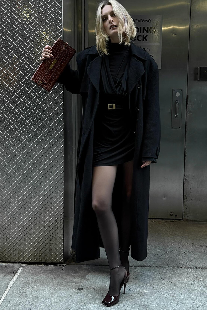 Shoe trends to wear with leggings, jeans, and skirts are shown in a street style photo of a woman standing in front of an elevator bank wearing a long black coat layered over a black blouse, black skinny belt, black mini skirt, and black sheer tights, styled with a red skinny clutch, and black patent leather peep-toe heels