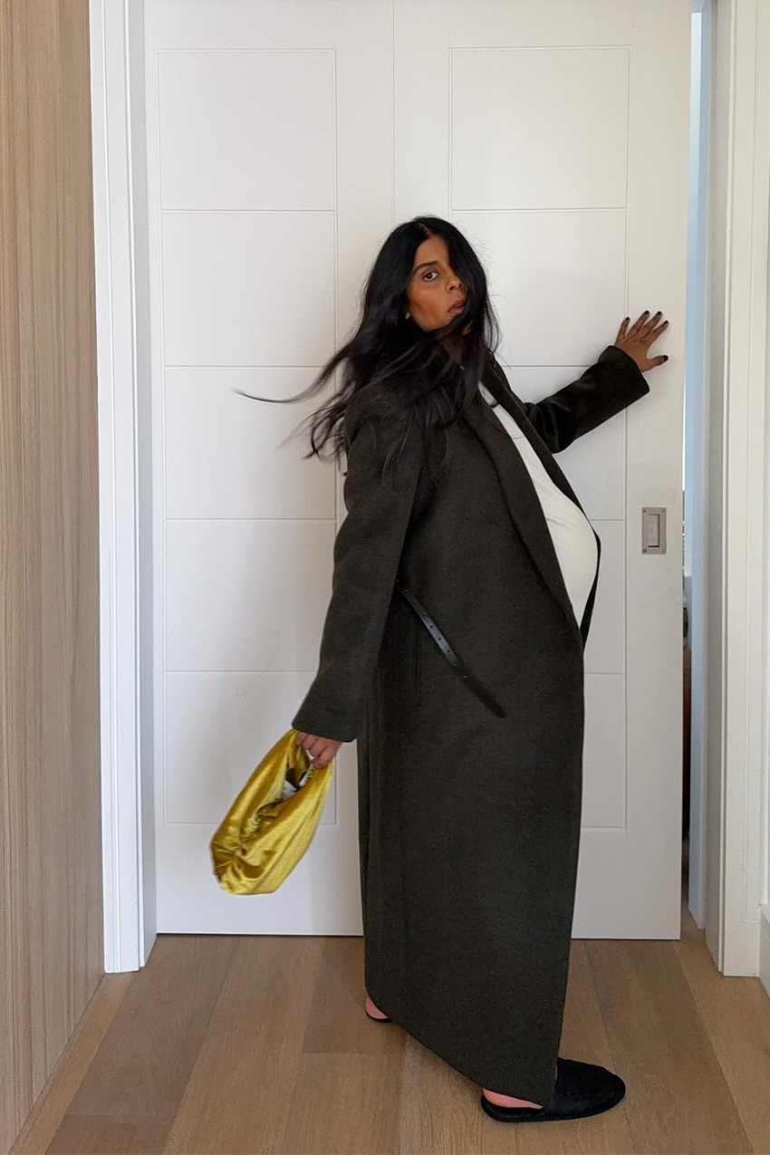 Shoe trends to wear with leggings, jeans, and skirts are shown in a photo of a woman standing inside her home in front of a door, wearing a long olive green maxi coat over a white maxi sweater dress styled with a yellow satin clutch bag and black slippers