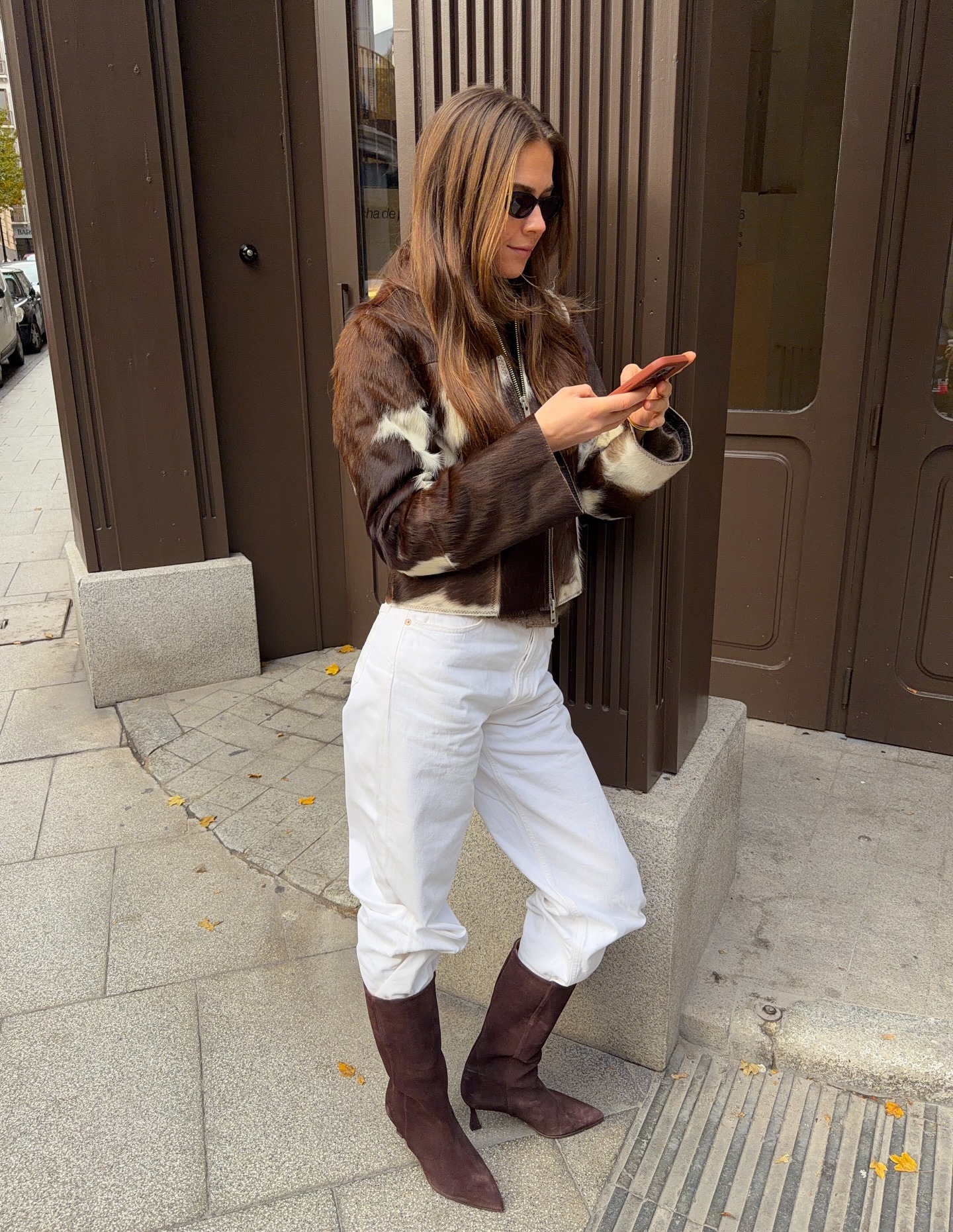 Woman wears a cow print pony hair jacket, white jeans, and suede boots while texting on the street.