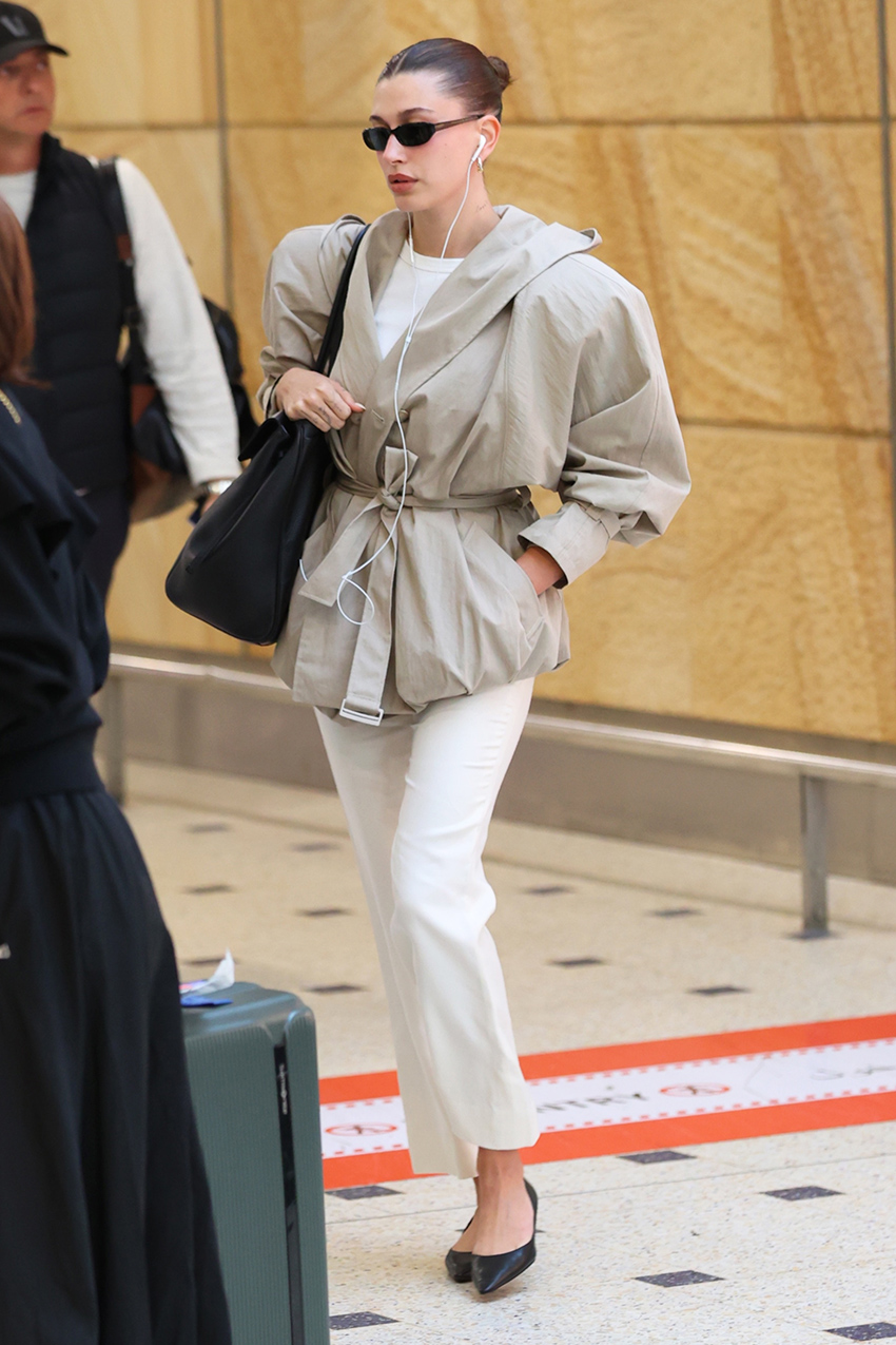 Hailey Bieber wearing a tan jacket, white pants, and kitten-heel pumps at the airport in Australia.