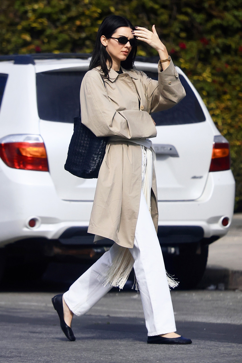 Kendall Jenner wearing a trench coat, scarf belt, white jeans, and black flats.