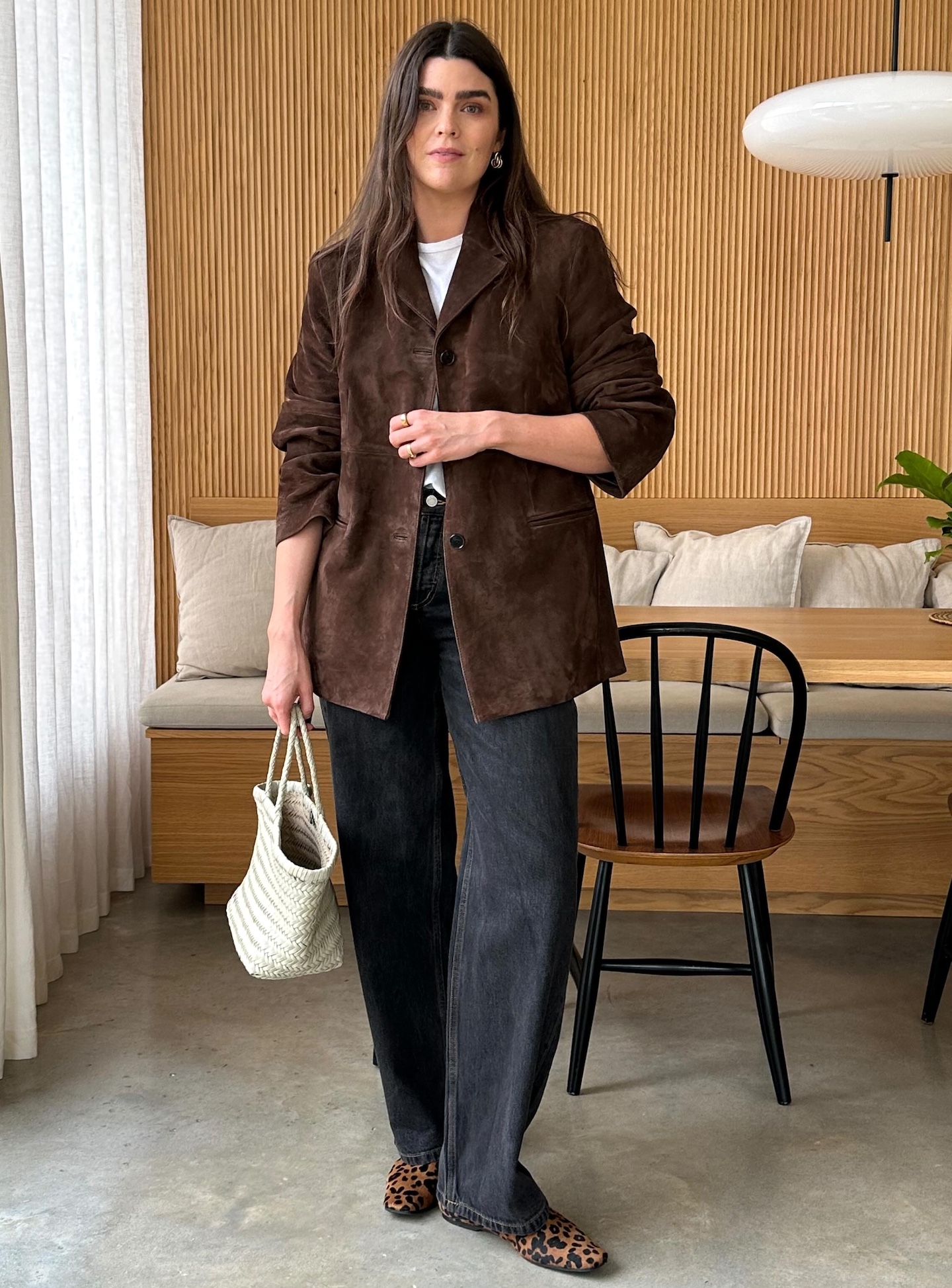 British style influencer Anna Howard poses in her London dining room wearing a brown suede jacket, white tee, black jeans, a woven tote bag, and leopard-print flats