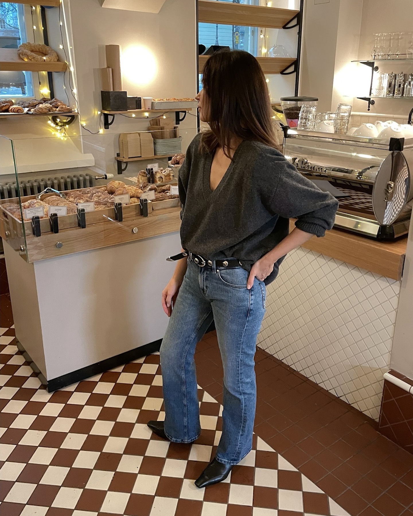 style influencer Johanna Piispa poses in a cafe wearing a gray deep V-neck sweater, studded belt, bootcut jeans, and black kitten-heel boots