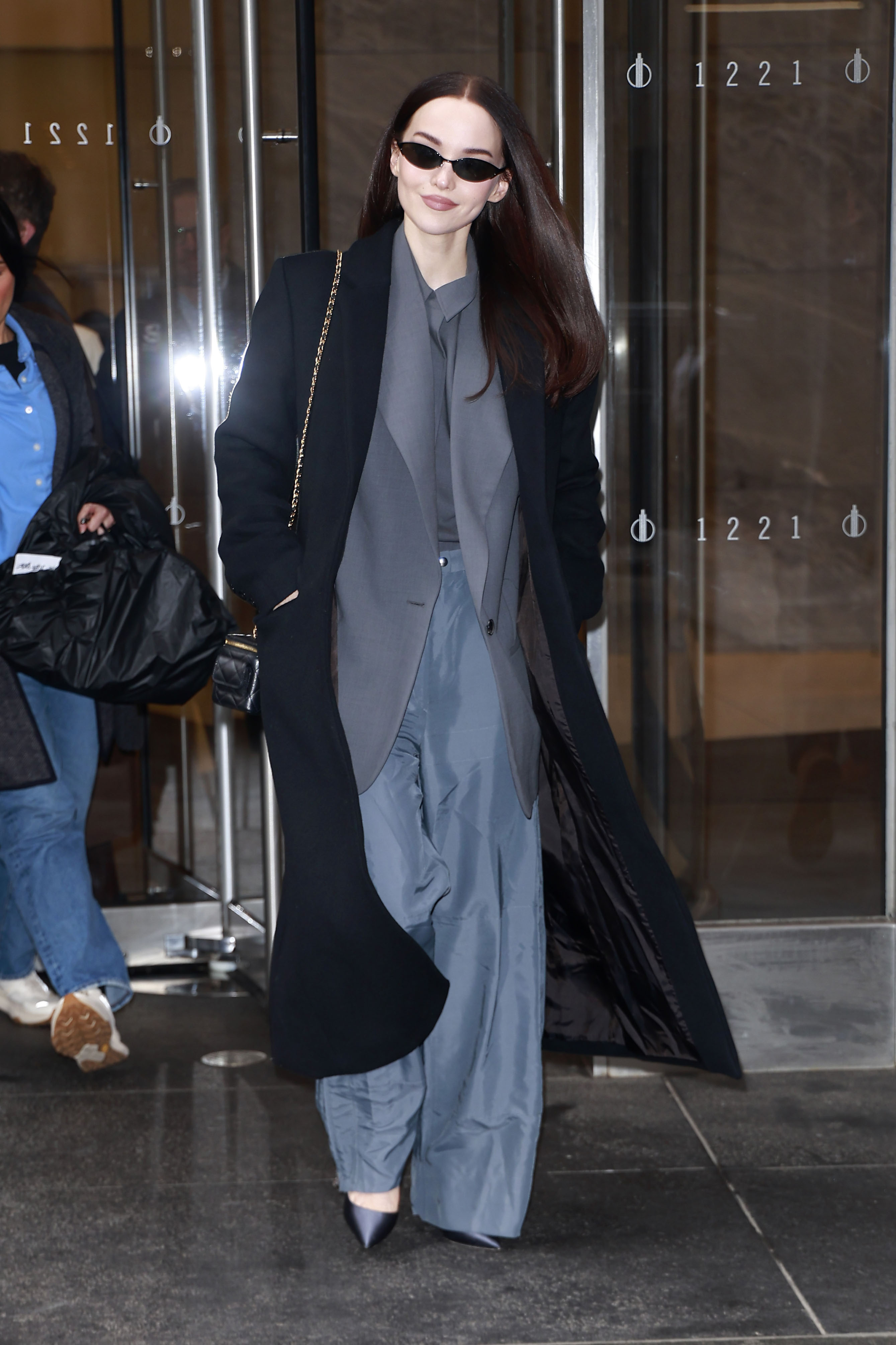 Dove Cameron wearing a black coat, gray blazer, gray pants, a black shoulder bag, and black heels.