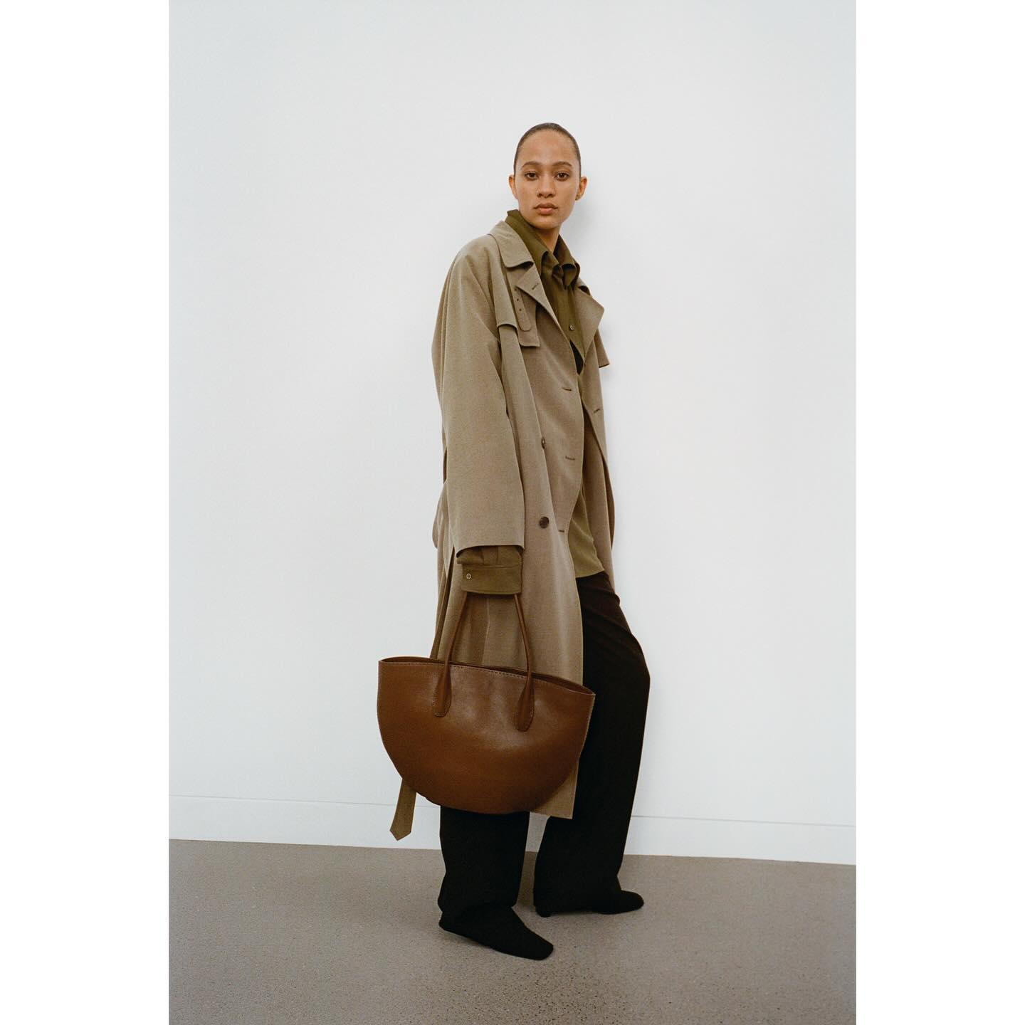 Camplaign image from The Row depicting a model wearing a brown trench coat, brown leather bag and black trousers.