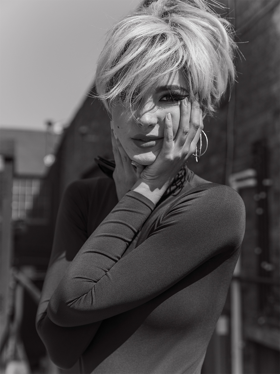 Black and white photo of actress Isabel May shot by Elias Tahan. She is wearing a short blonde wig with a mod-inspired cat eye liner, a long sleeve T-shirt, and hoop earrings. Her hands are framing her face.