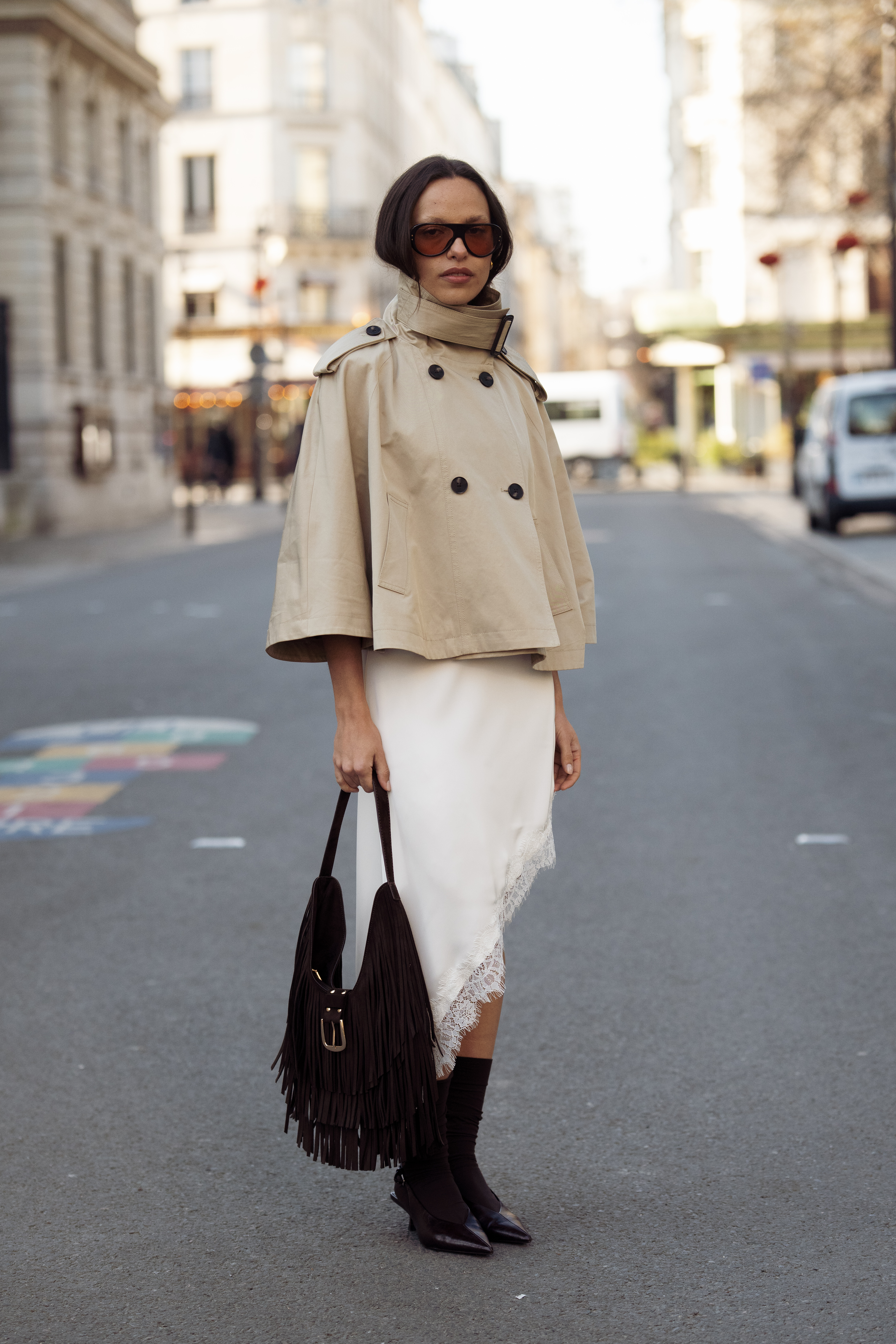 PARIS, FRANCE - MARCH 04: A guest wears a beige cropped trench coat with wide sleeves and a high buckled collar over a cream asymmetric satin slip dress with lace trim, brown oversized sunglasses, carries a dark brown suede fringe shoulder bag, black socks and black pointed kitten heel pumps outside Courreges show during Day Three of Paris Fashion Week Womenswear Fall/Winter 2026/2027 on March 04, 2026 in Paris, France. (Photo by Raimonda Kulikauskiene/Getty Images)