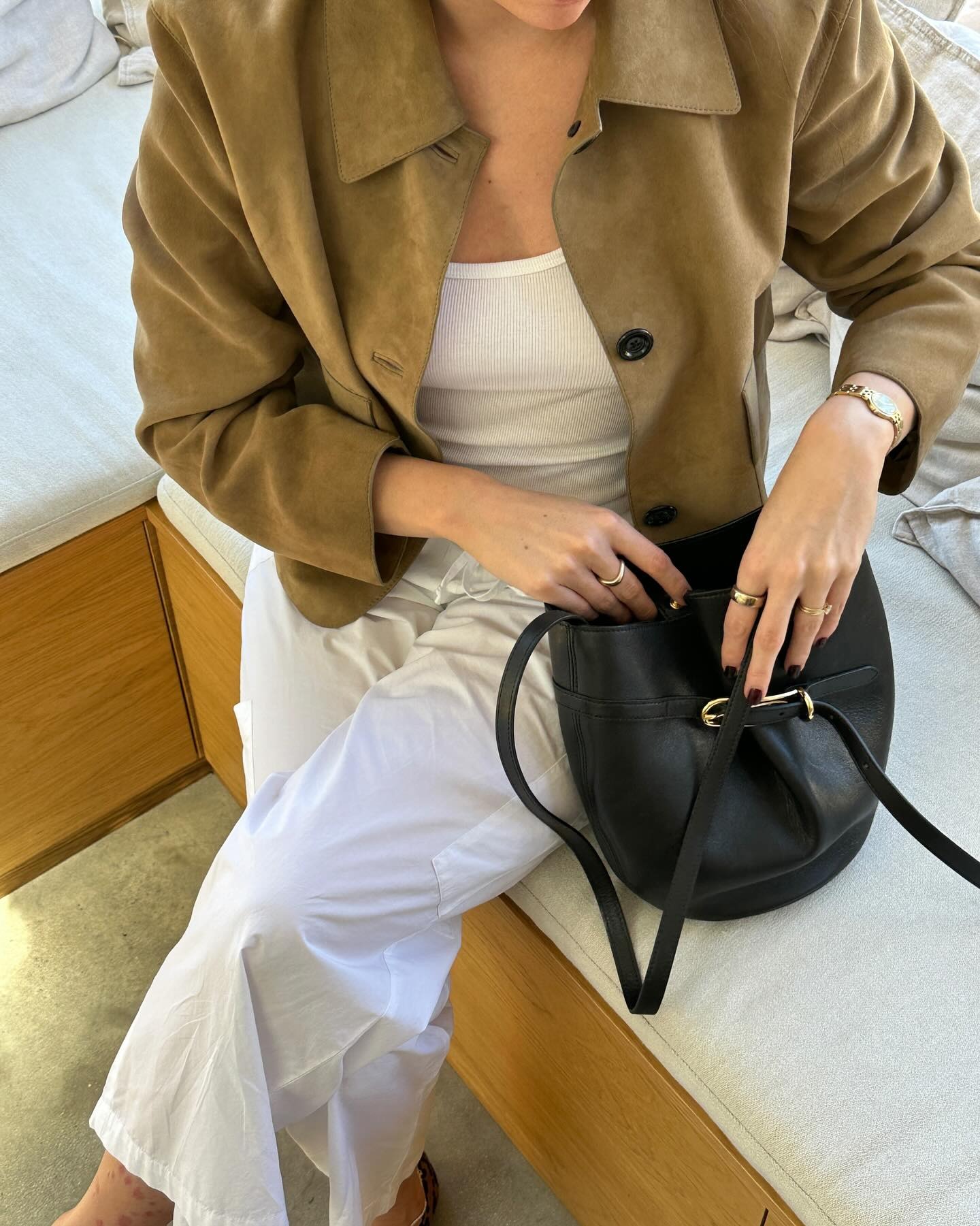 British style influencer Anna Newton wearing a tan suede jacket, a white ribbed shirt, white relaxed pants, and a black Little Liffner buckled bucket bag