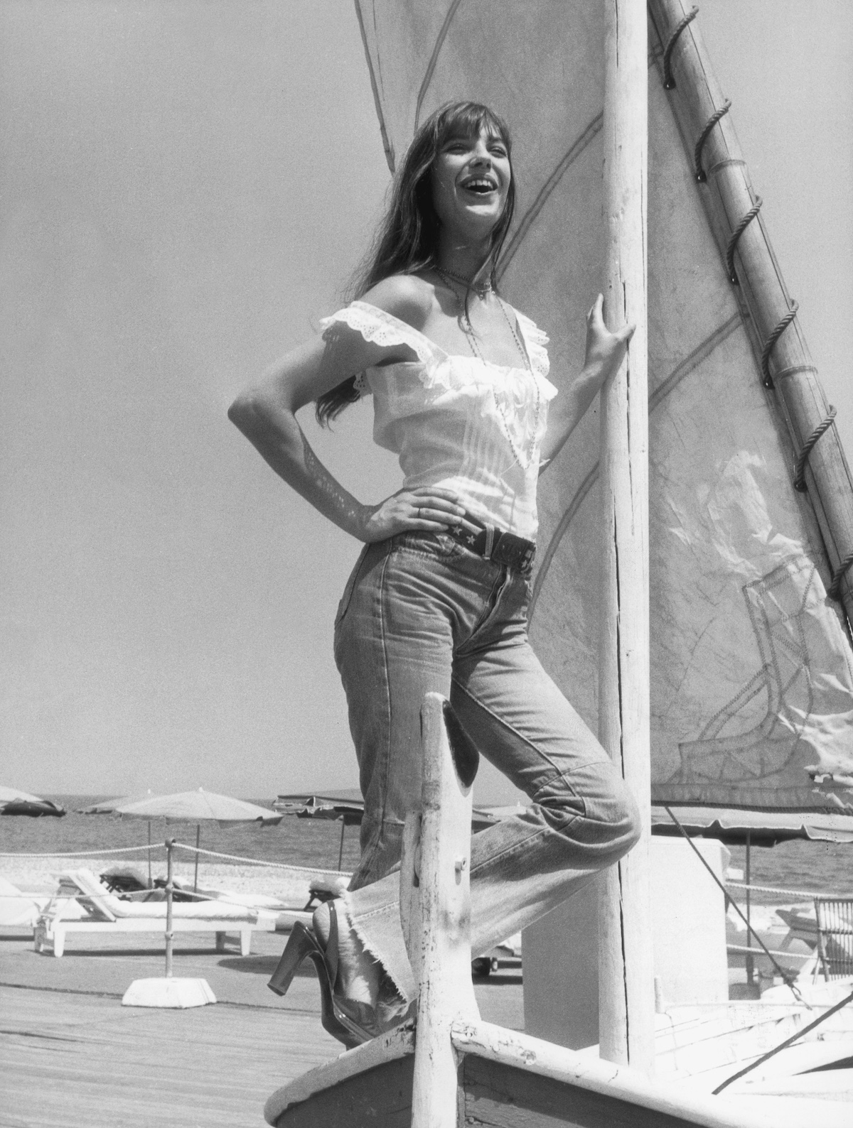 Jane Birkin posing on a sail boat wearing a white ruffle top, belt, flared raw-hem jeans, and platform slingback heels