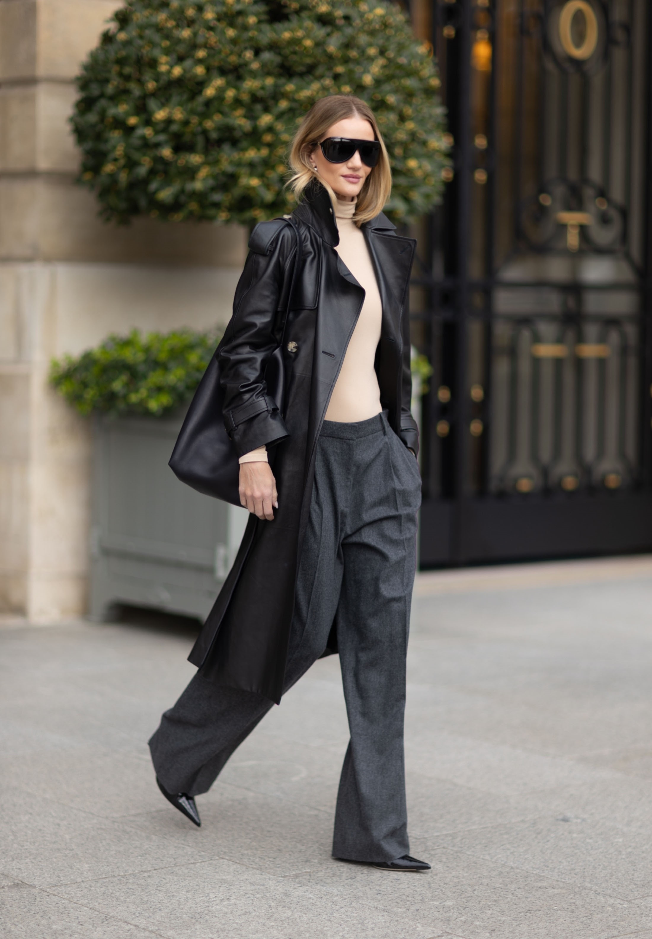 Rosie Huntington steps outside in Paris wearing grey trousers and a black leather trench coat.