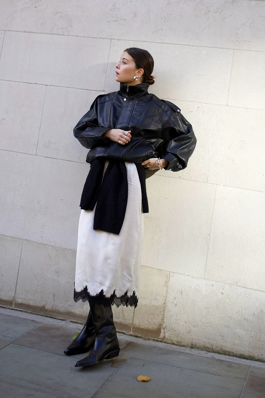 Layering staples for spring 2026 is shown in a street style photo with a woman standing outside while wearing silver earrings, a leather bomber jacket, a white slip skirt with black lace trim, a black cardigan tied around the waist, and black flat knee-high boots