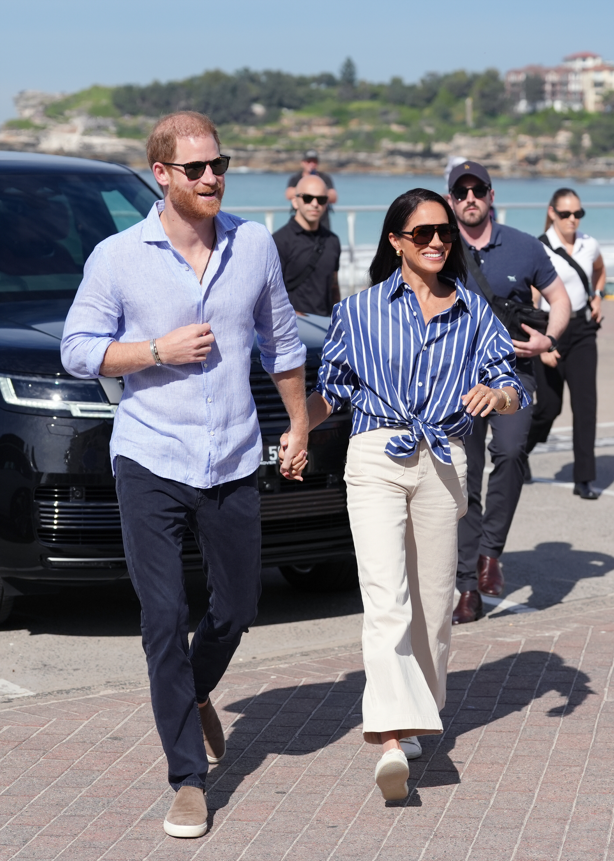 Meghan Markle holds hands with Prince Harry wearing white cropped jeans with white trainers and a blue striped shirt.