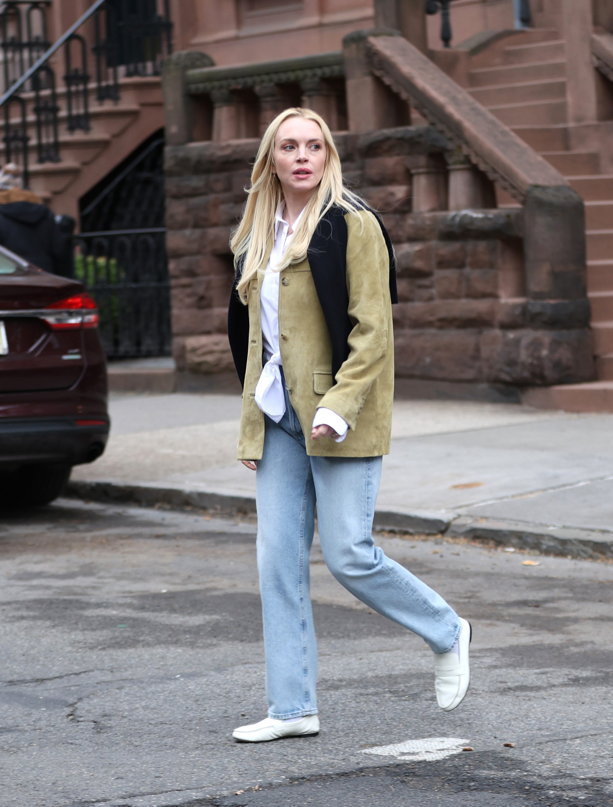 Lindsay Lohan wears a suede jacket, white button-down shirt, black sweater, light-wash jeans, and white loafers.