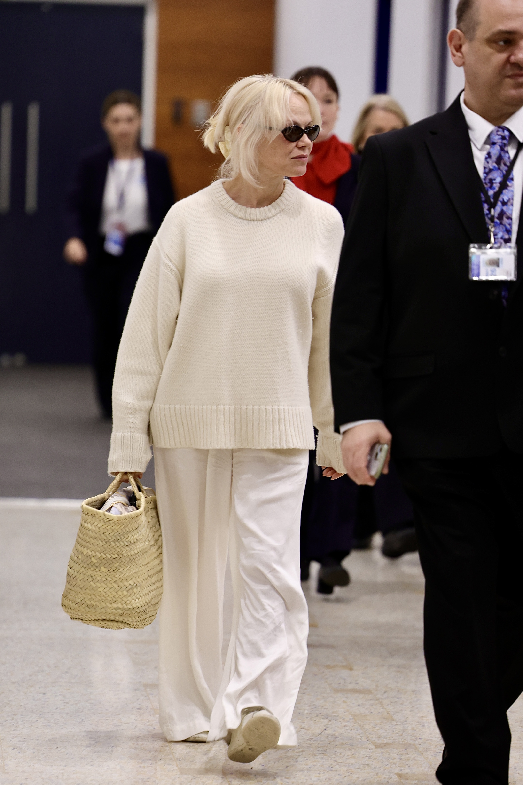 Pamela Anderson walks through the airport wearing white trainers with a wicker bag, a white jumper and white trousers.