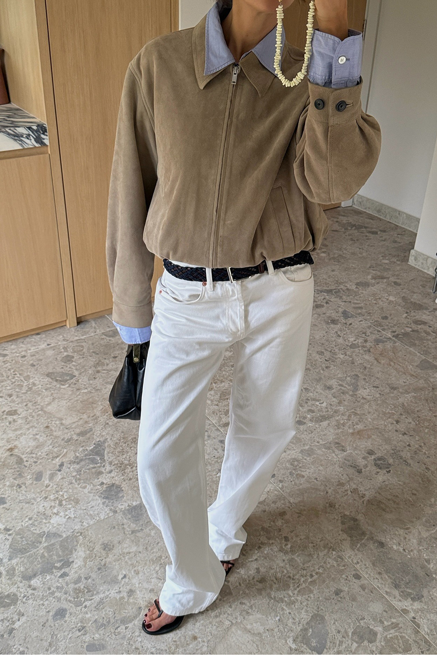 a woman wearing a tan suede jacket, white jeans, and black flip flops