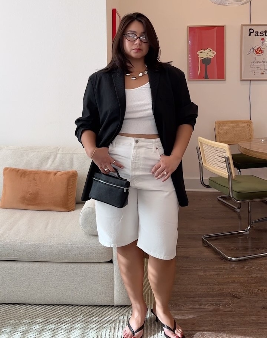 style influencer Marina Torres poses in her living room wearing oval optical eyeglasses, a black blazer, white tank top, white denim Bermuda shorts, mini black bag, and black kitten-heel flip-flips