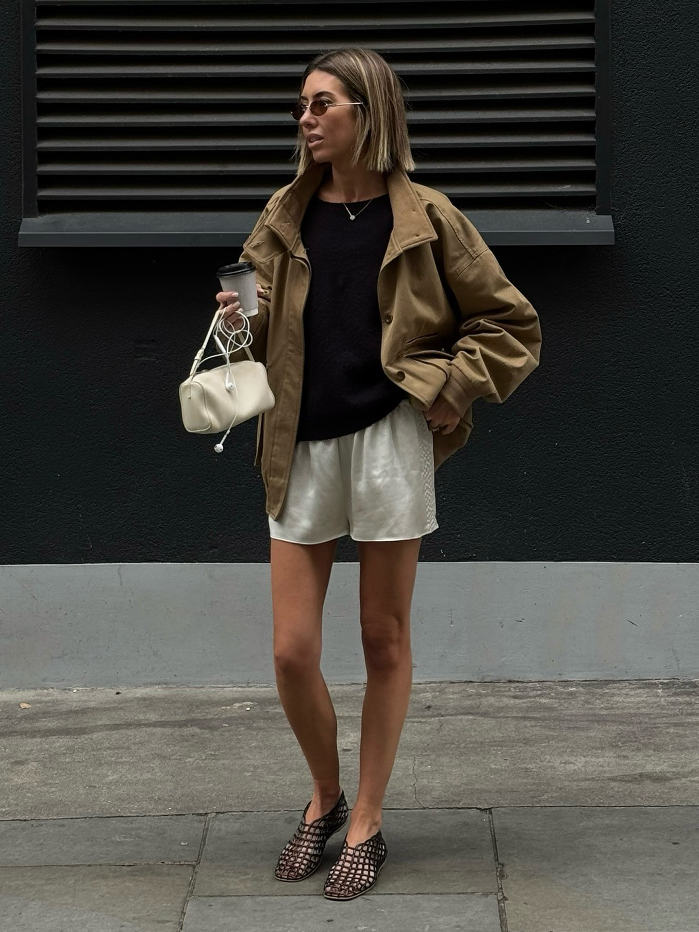 British style influencer Hannah Lewis poses in London wearing a tan jacket, black top, ivory silk shorts, mini white bag, and black jelly flats