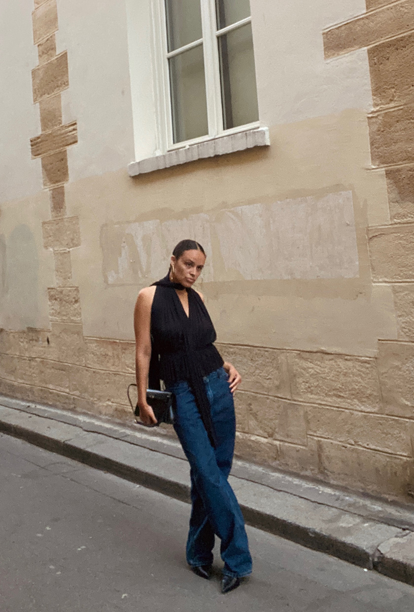 An editor standing on a street wearing a black halter top with baggy jeans, black pointed heels, and a black shoulder bag.