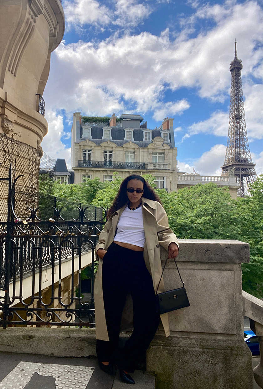 An editor standing on a staircase in front of the Eiffel Tower wearing a trench coat with a T-shirt, low-rise trousers, and pointed flats.