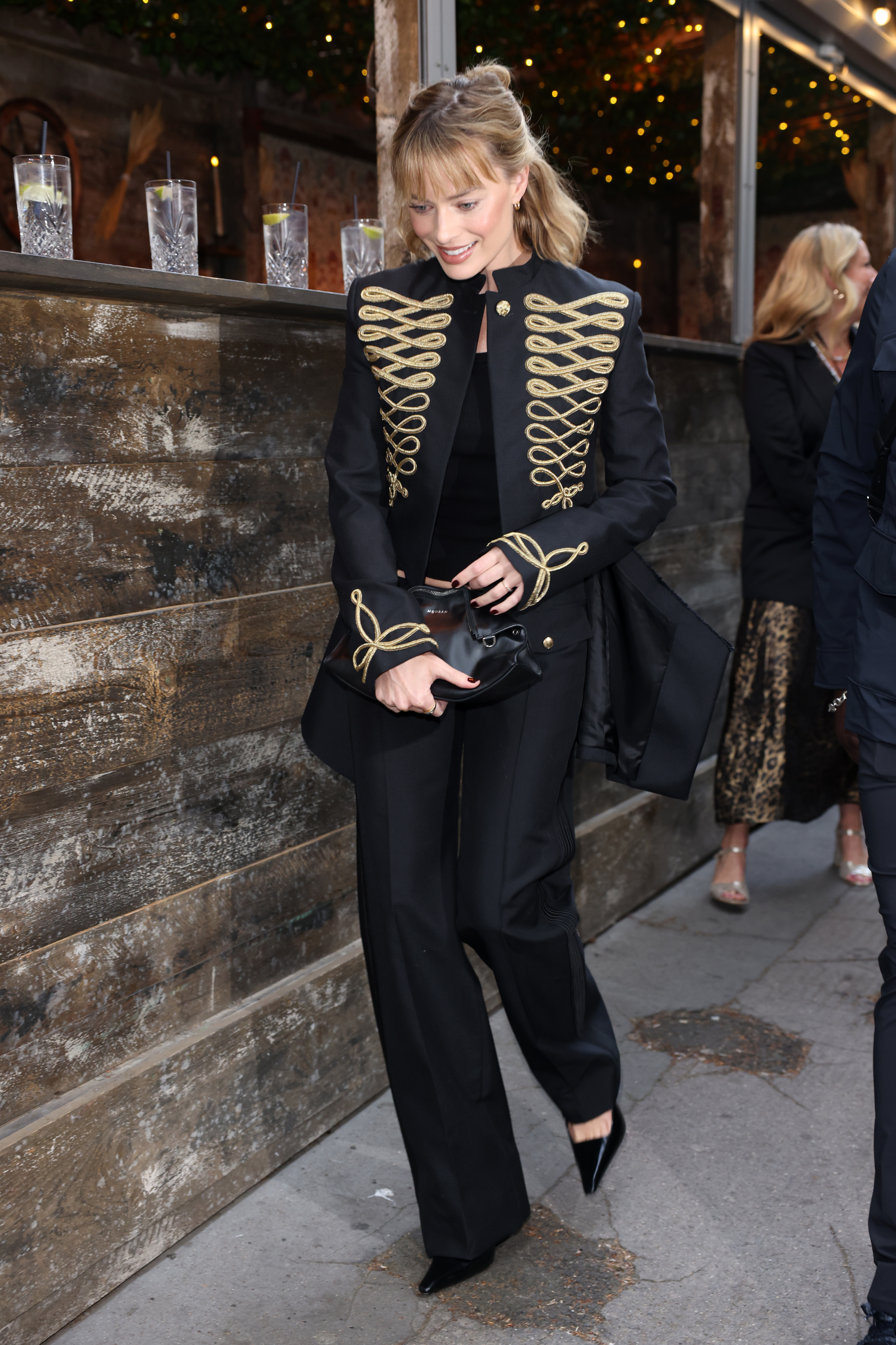 Margot robbie wears a black and hold military jacket, low-rise black trousers, black pumps, and a black clutch.
