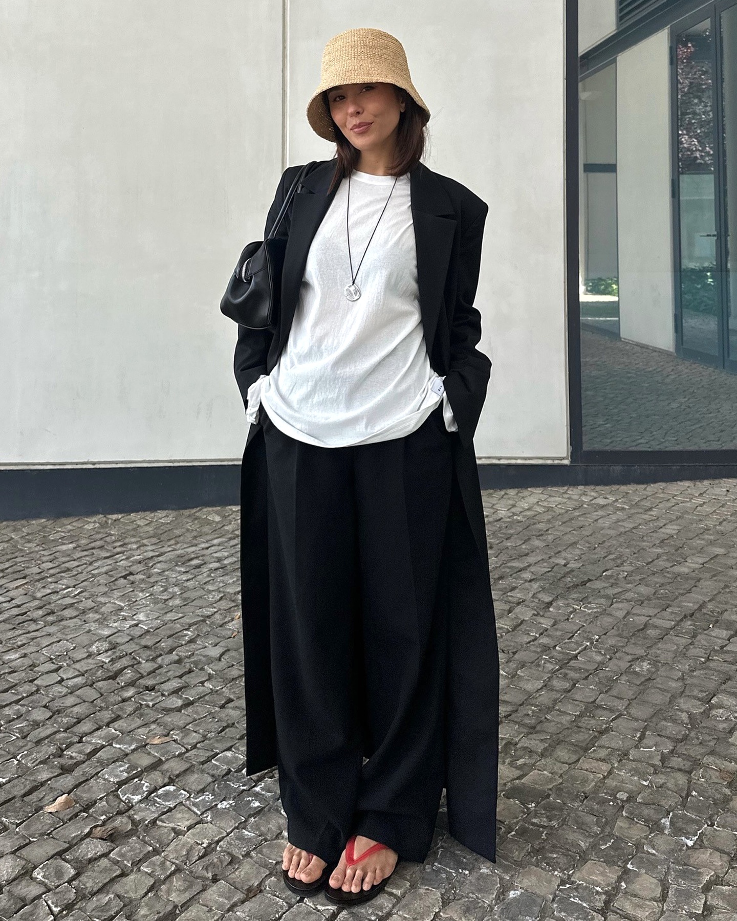 style influencer Debora Rosa poses on a cobblestone street in Lisbon, Portugal wearing a bucket hat, black maxi coat, white T-shirt, long pendant cord necklace, black pants, and red flip-flops
