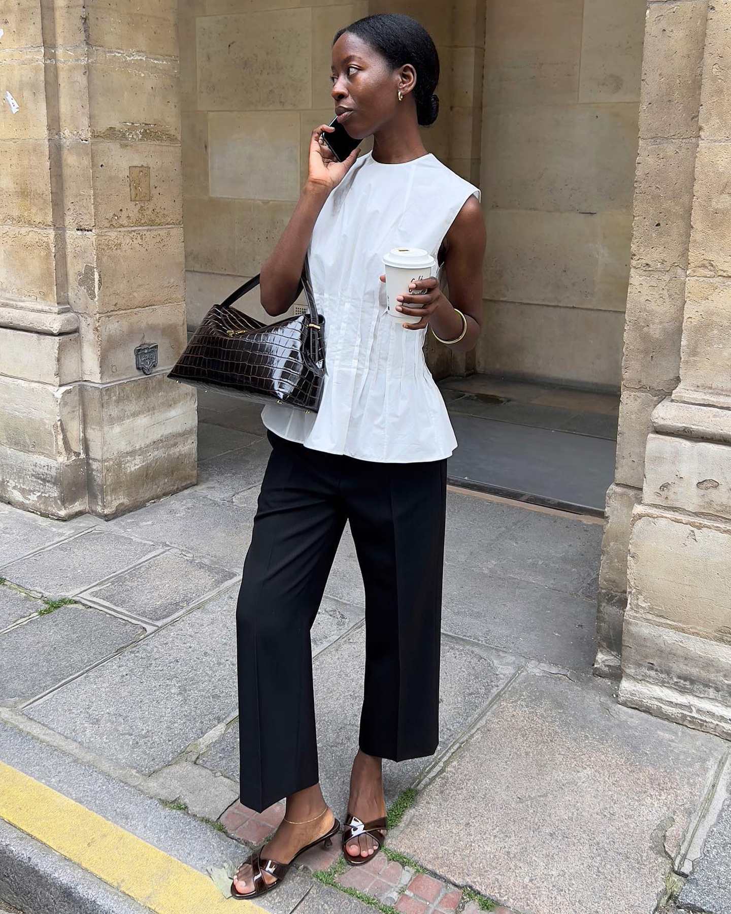 influencer and stylish Sylvie Mus poses on her phone and holding a coffee, wearing a white sleeveless pleated top, a brown croc-embossed bag, cropped black pants, and brown mule kitten-heel sandals