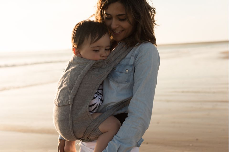 Junge Mutter spaziert mit schlafendem Kind in der Babytrage am Strand.