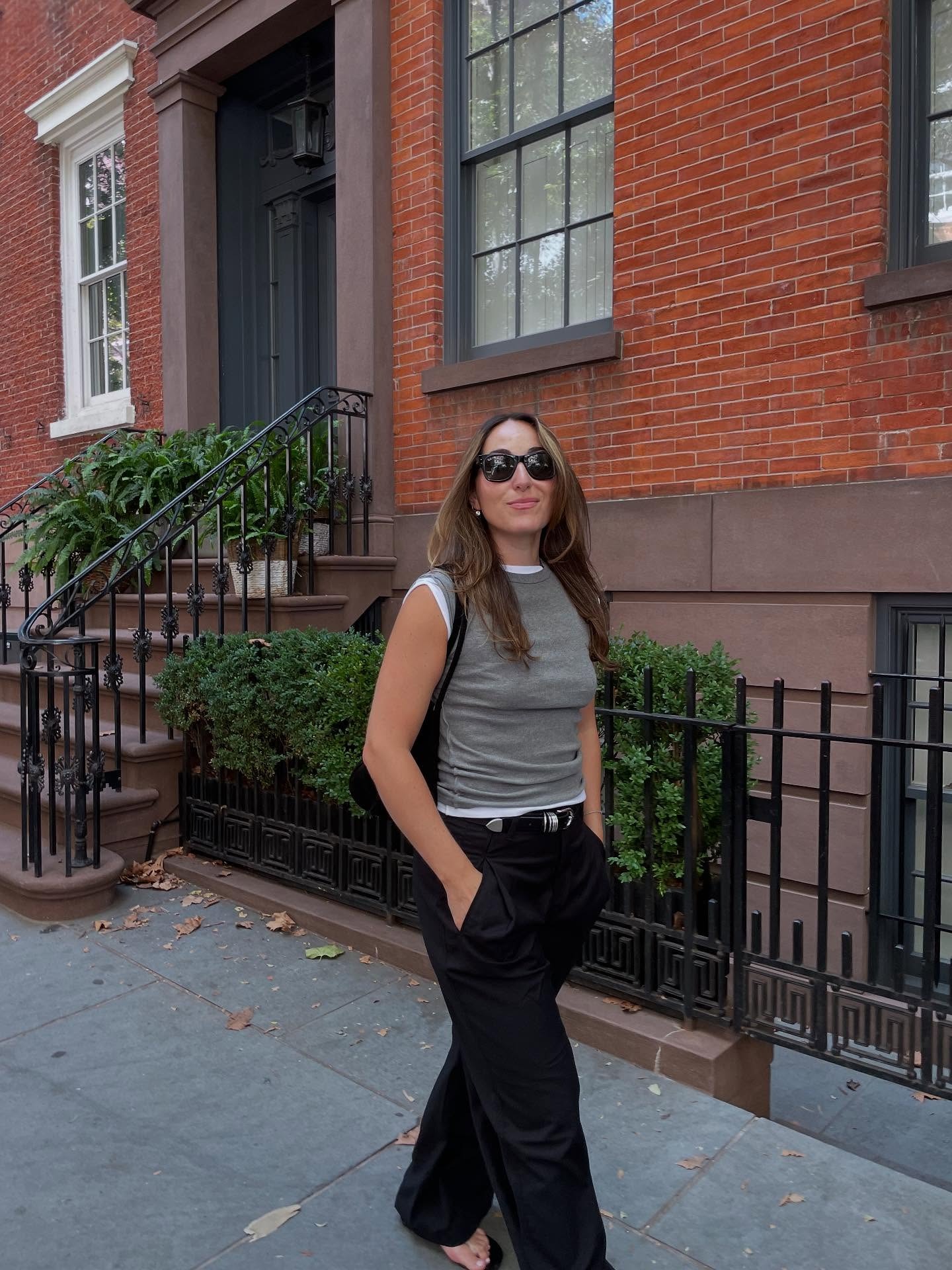 Anna wears two tees layered over each other. One is gray and one is white. She also wears black trousers and thong sandals.