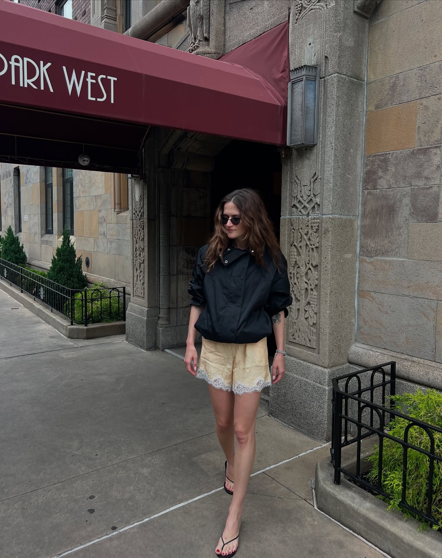 Eliza wears funnel neck black windbreaker, lace shorts, and flip flops.
