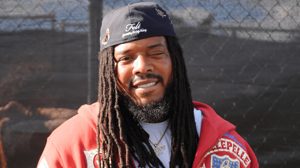 A smiling man with dreadlocks wearing a red jacket and a baseball cap with the text "Fell Clothing Company".
