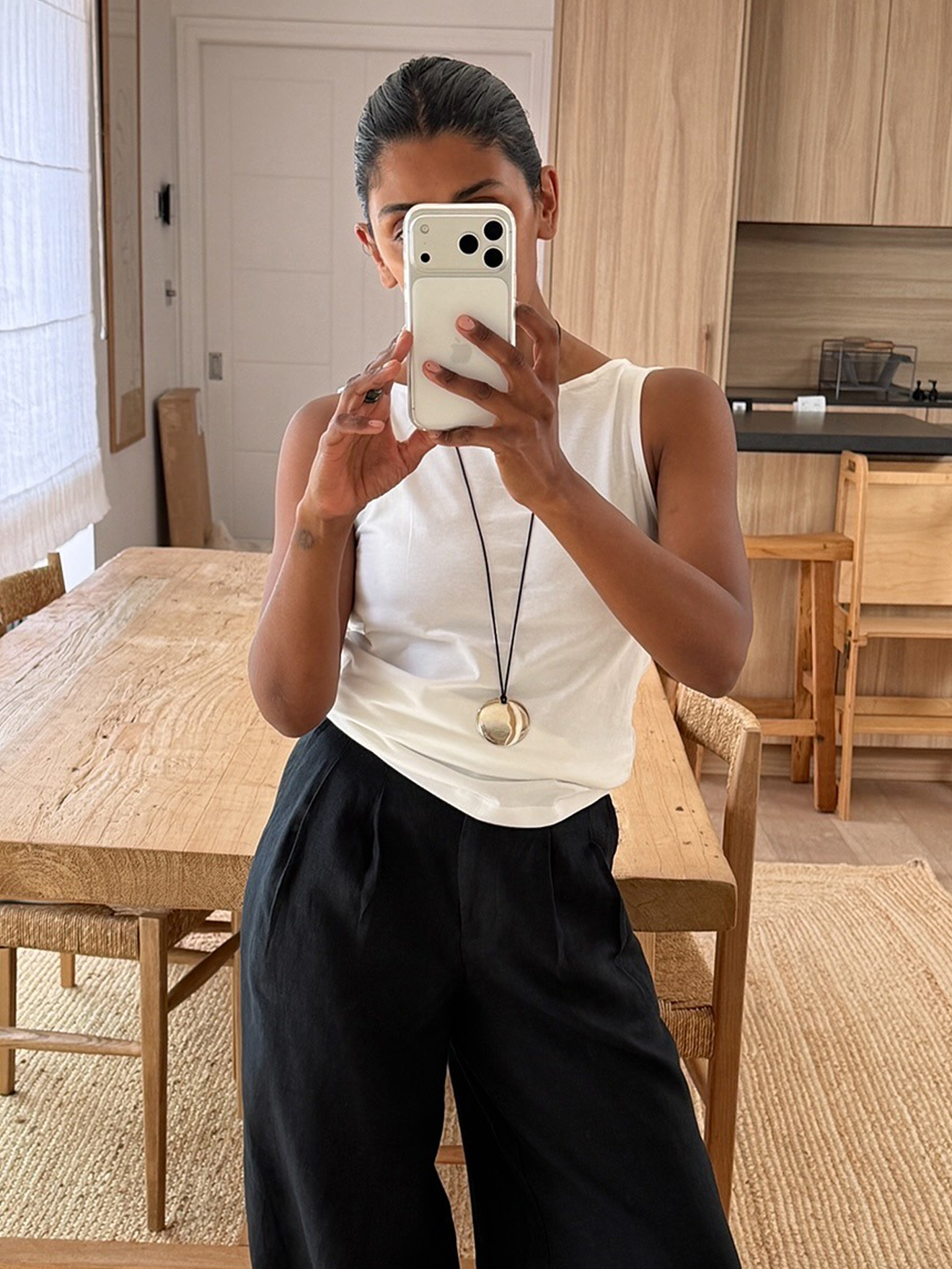British style influencer Monikh Dale poses for a mirror selfie in her London home wearing a white boatneck tank top, long cord necklace with pendant, and black relaxed pants