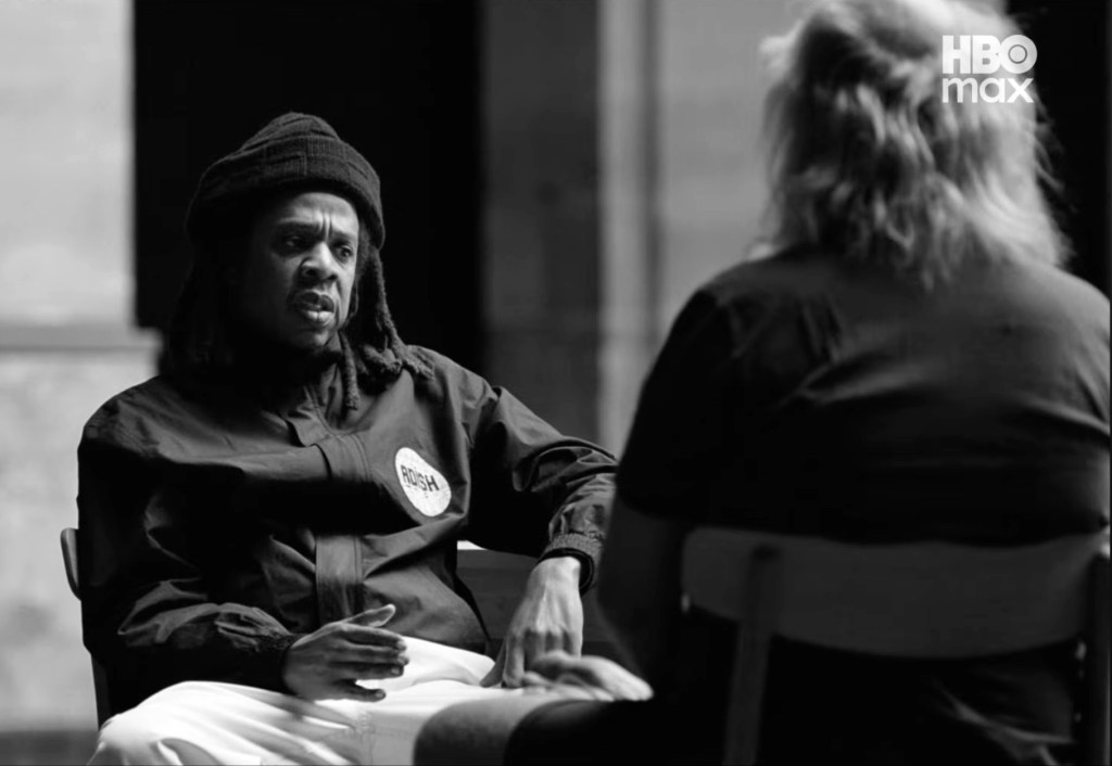 A Black man wearing a "PUSH" jacket sits on a bench, speaking to a woman in a dark jacket.