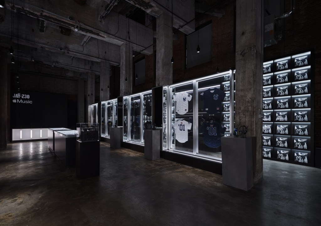 A dark, industrial-style room with shelves displaying various sports jerseys and memorabilia behind glass panels. The room is illuminated by bright lighting, creating a dramatic, museum-like atmosphere.