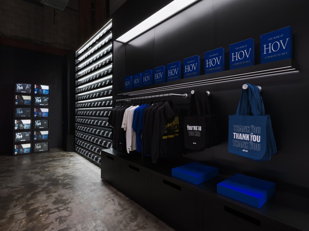 Shelves of books titled "The Book of HOV" in a dark retail space, with clothing and tote bags on display.