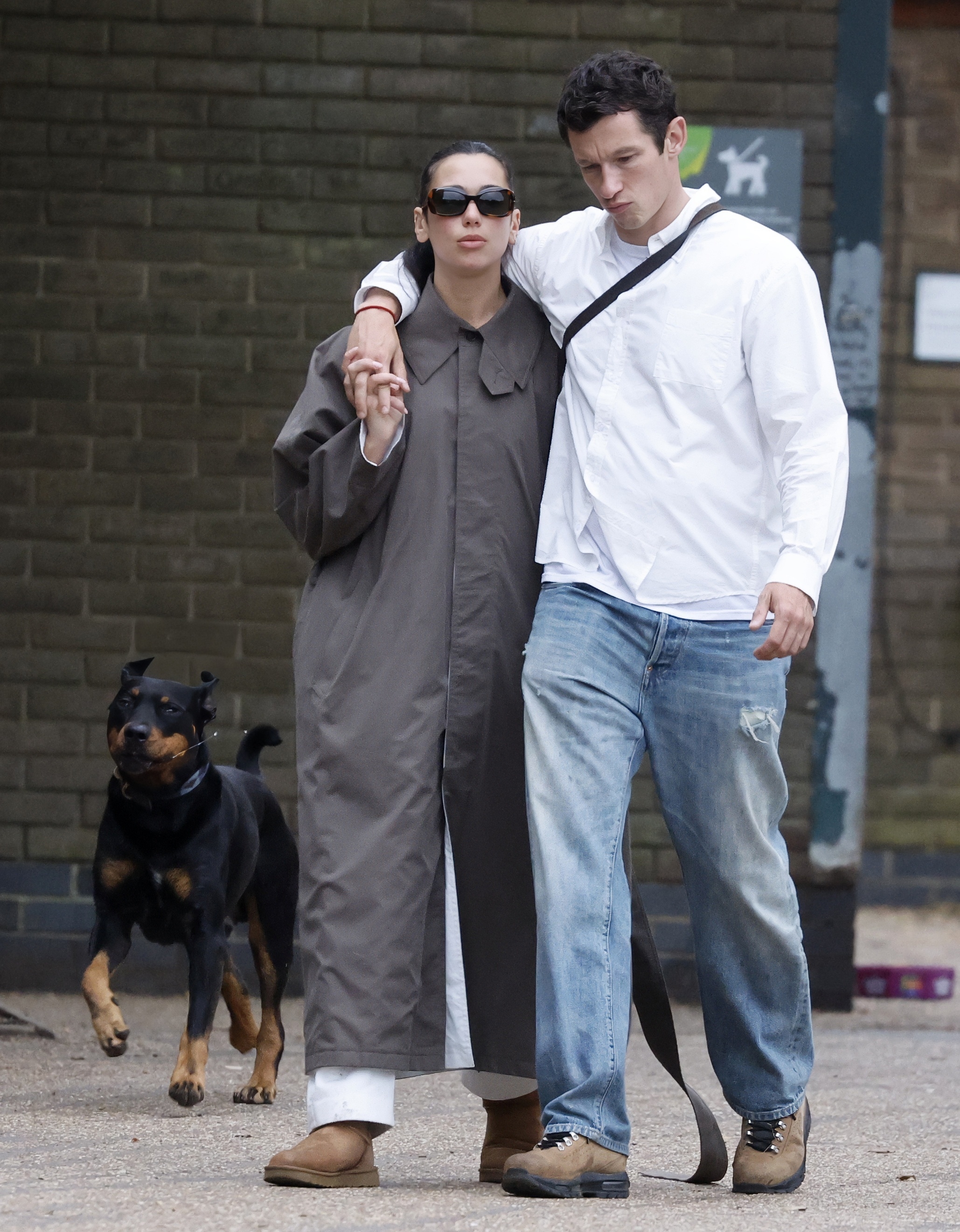 A photo of Dua Lipa and Callum Turner after their London wedding ceremony walking around the city together. Dua is wearing a gray, long trench coat, white jeans, and chestnut Ugg boots.