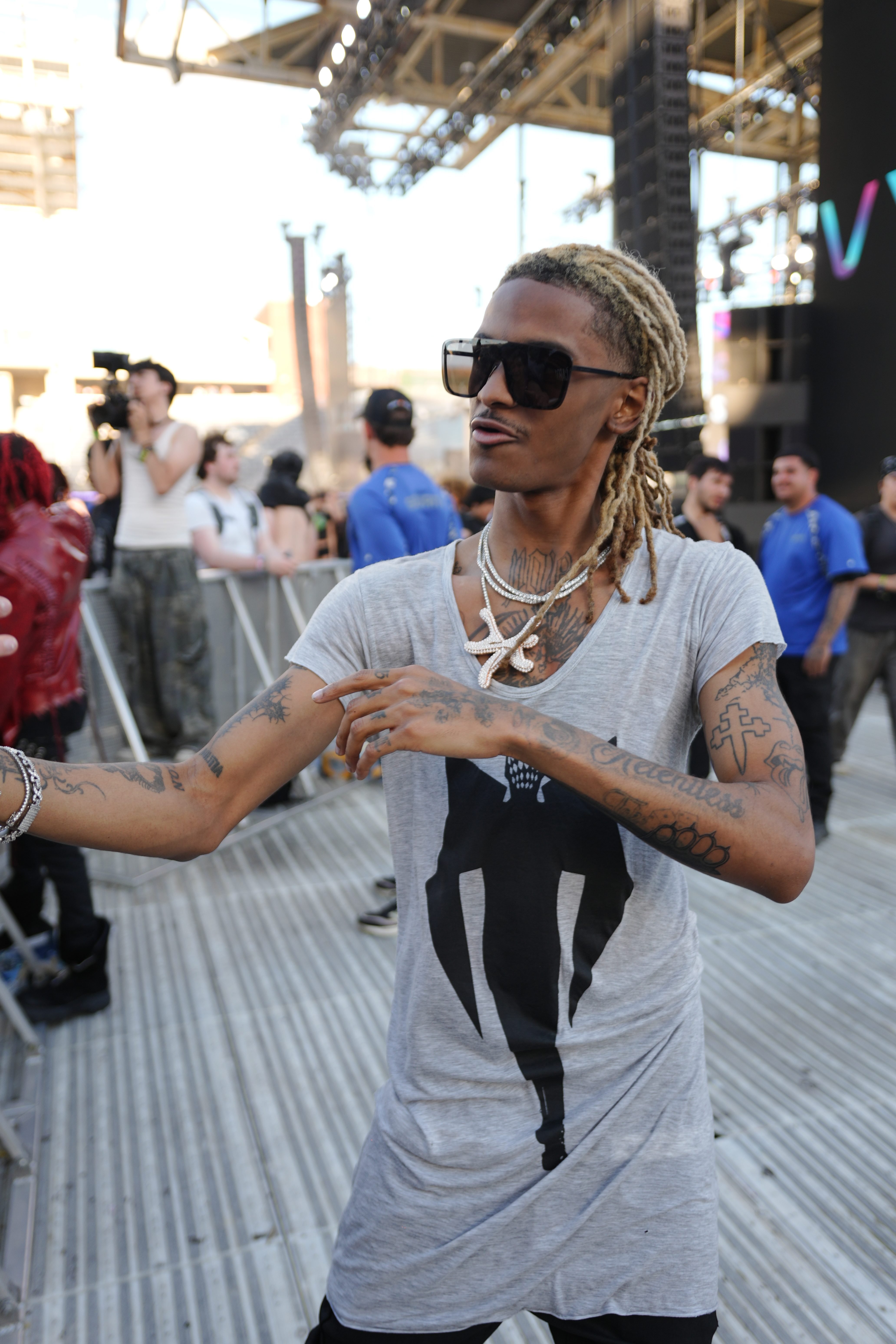 A person with dreadlocks and tattoos wearing sunglasses and a gray t-shirt with a graphic design, standing in a crowd at an event or concert.