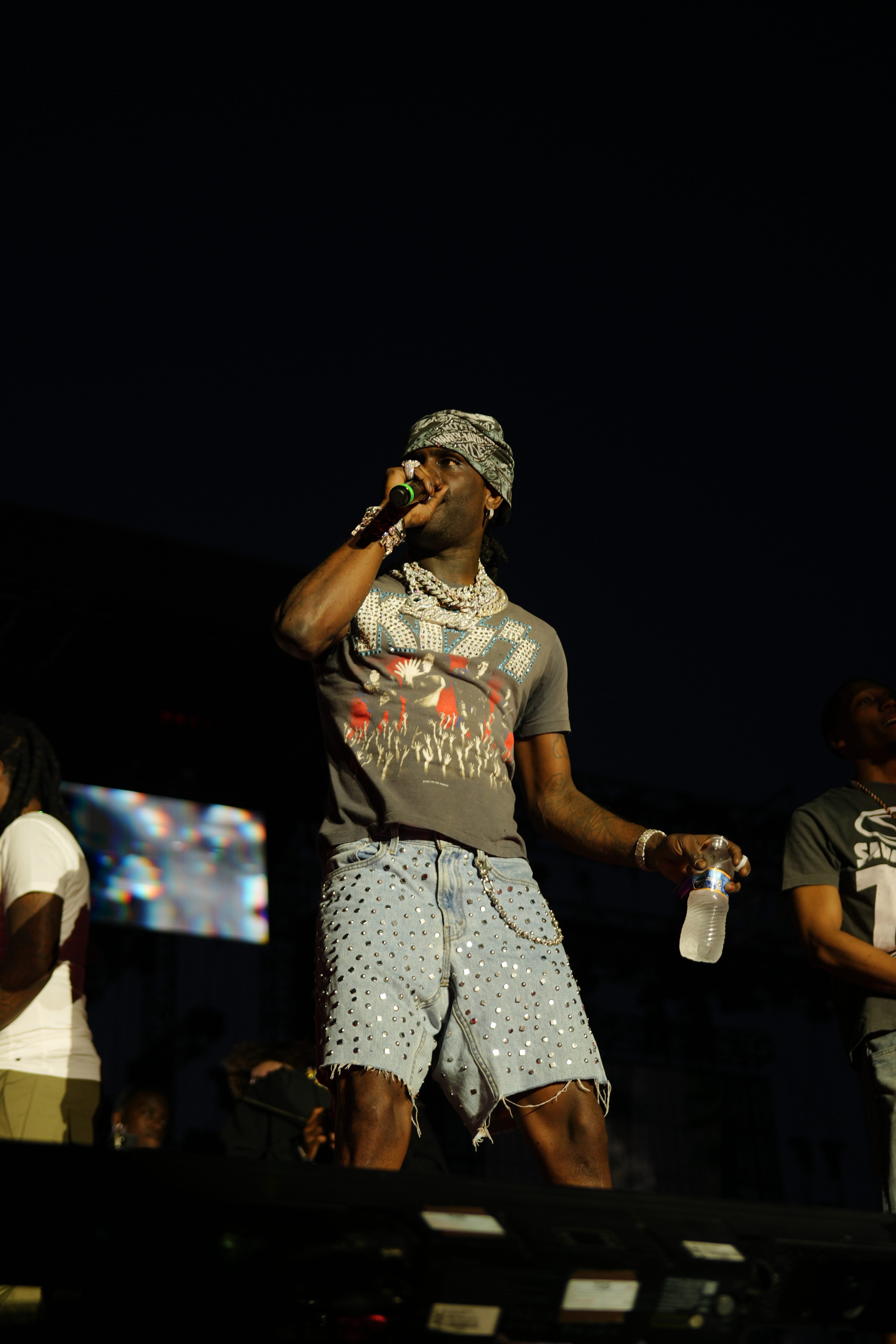 A performer wearing a patterned shirt and hat holding a microphone on stage in front of a dark background.