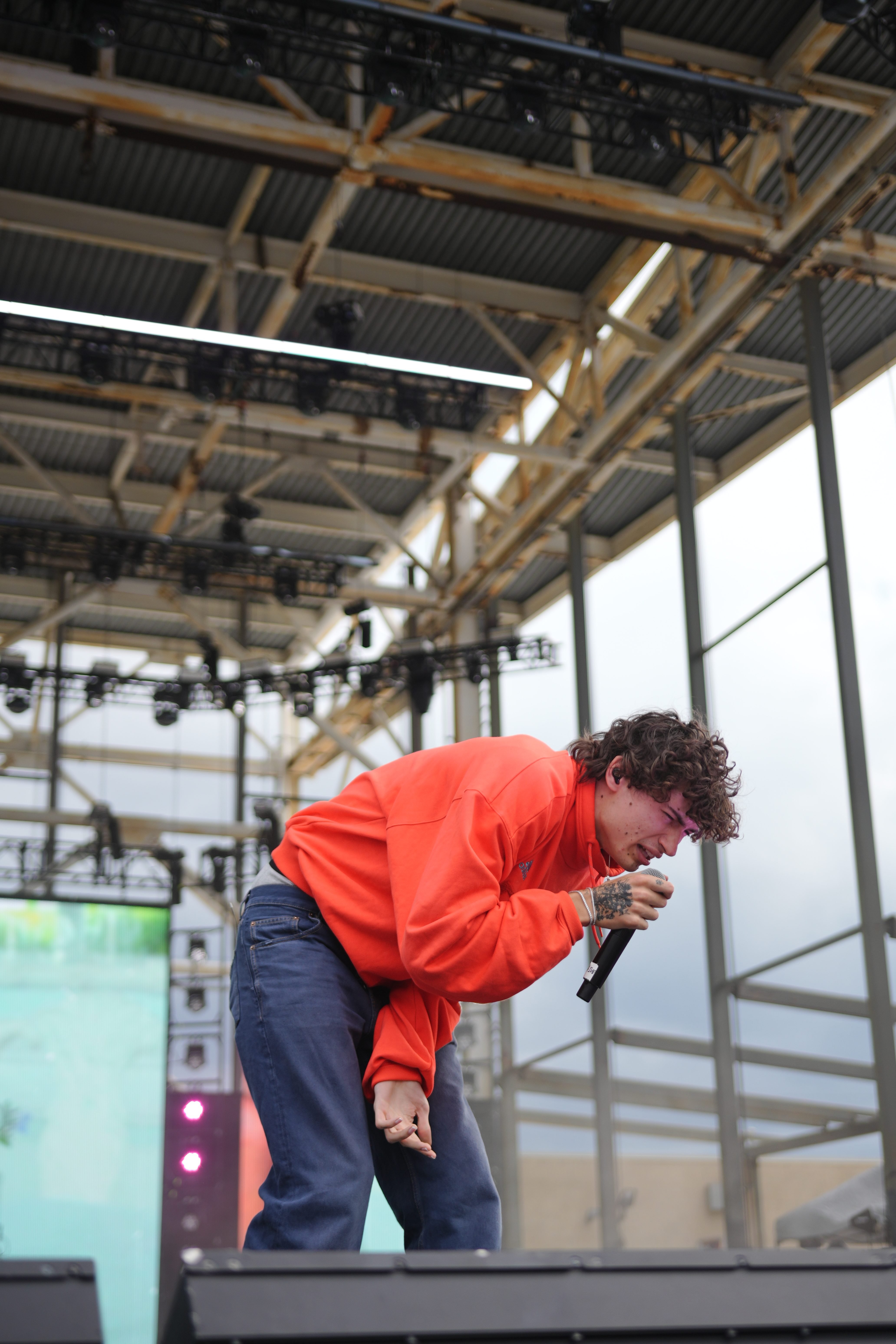 A person in a bright orange jacket performing on a stage with lighting equipment and scaffolding visible in the background.
