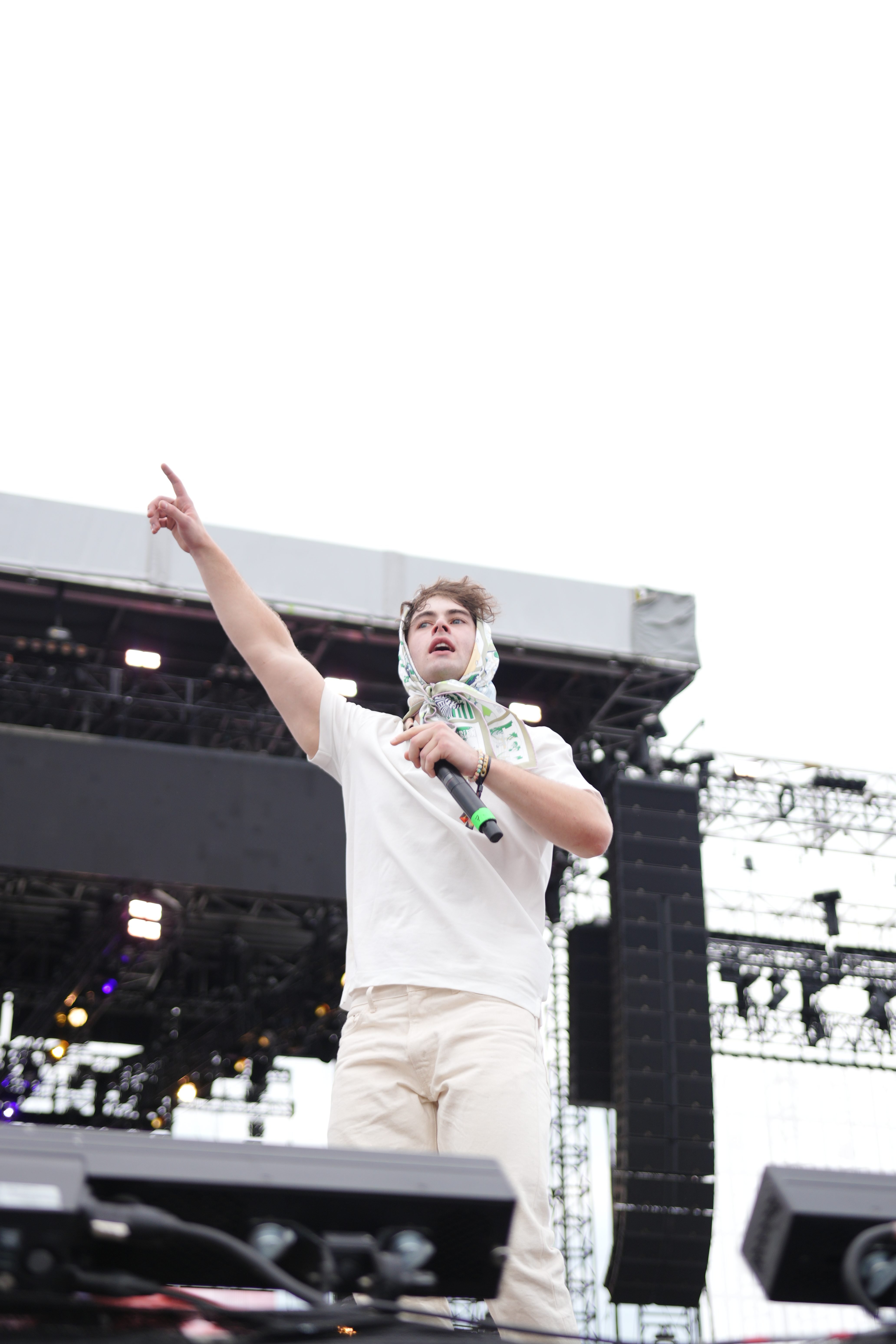 A person in a white shirt and scarf gesturing on a stage with lighting equipment and structures in the background.