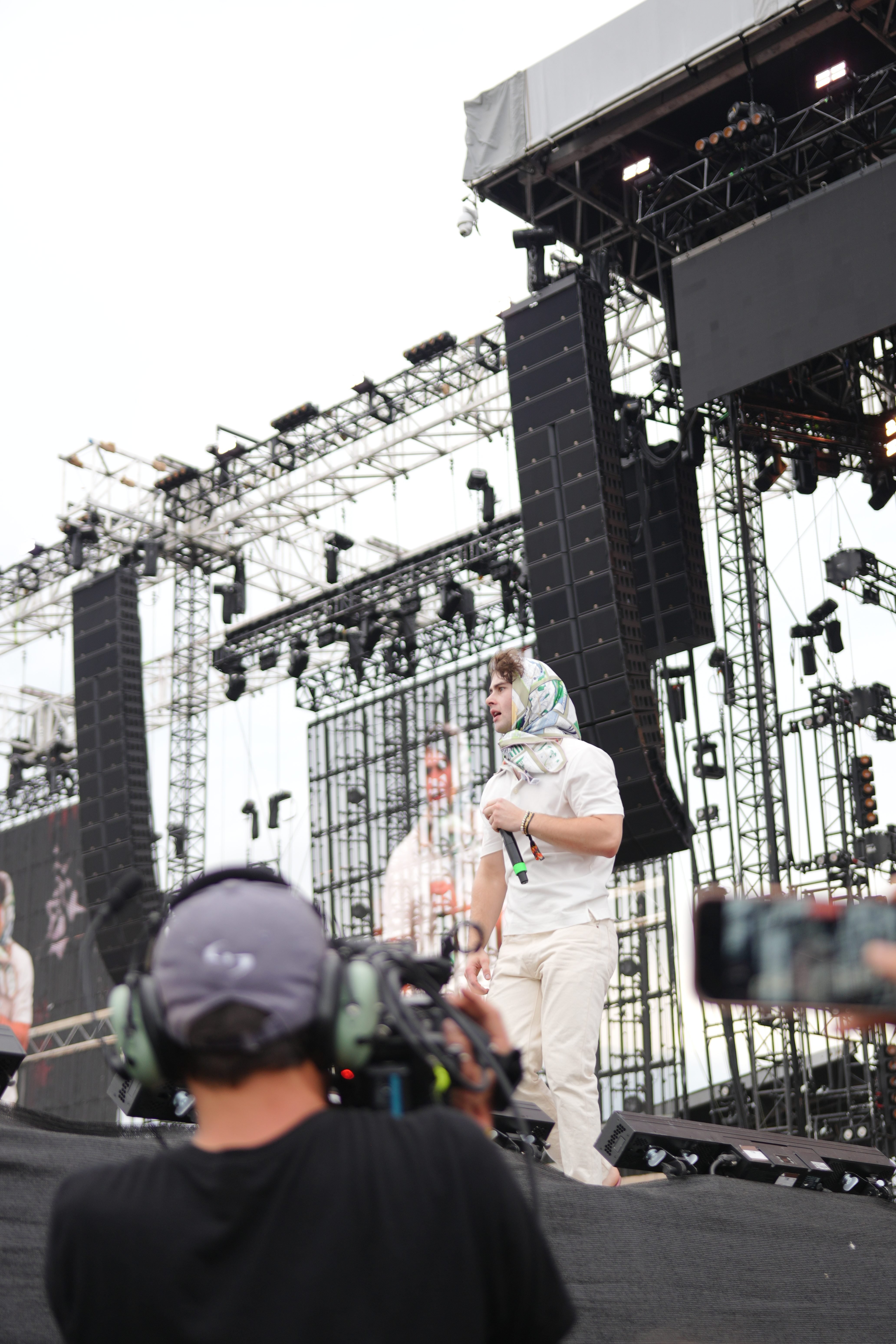 A person in a white outfit stands on a stage with lighting equipment and scaffolding visible in the background.