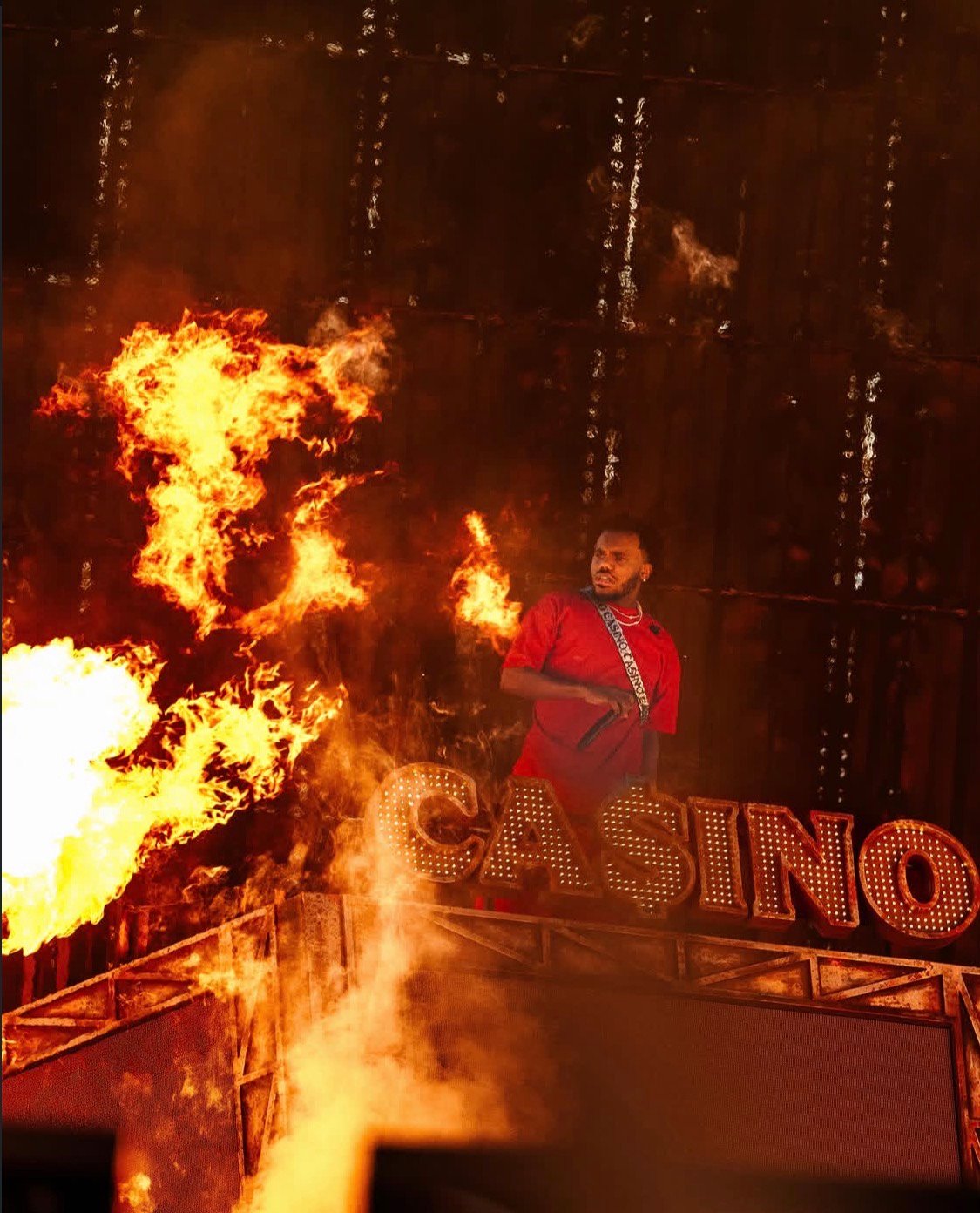 A person in a red costume stands in front of a large flaming "CASINO" sign, surrounded by intense flames and smoke.