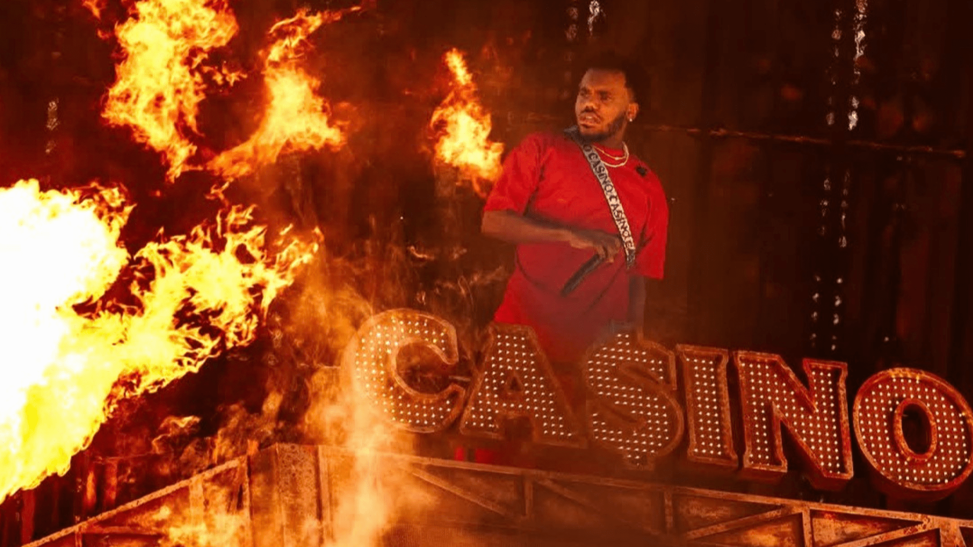 A person in a red costume stands on a platform surrounded by flames, with the word "CASINO" illuminated in large letters behind them.
