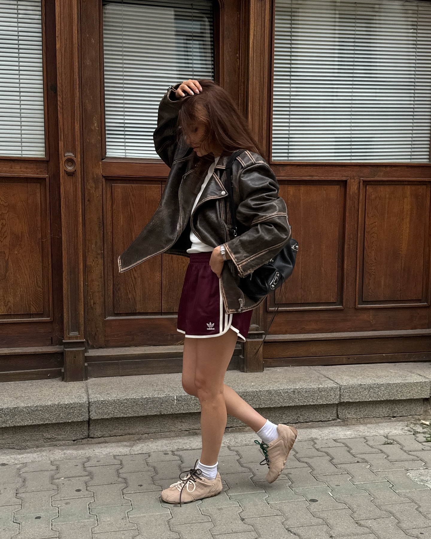 An image of an influencer wearing a brown leather jacket, maroon sports shorts and beige slim-sole trainers in Europe