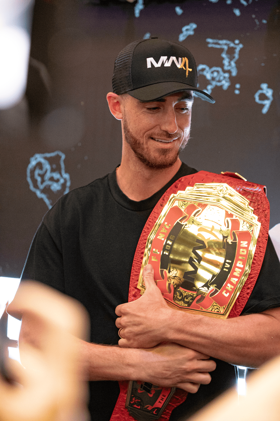 A man wearing a black MAVL trucker hat and holding a championship belt with the text "CHAMPION" on it.