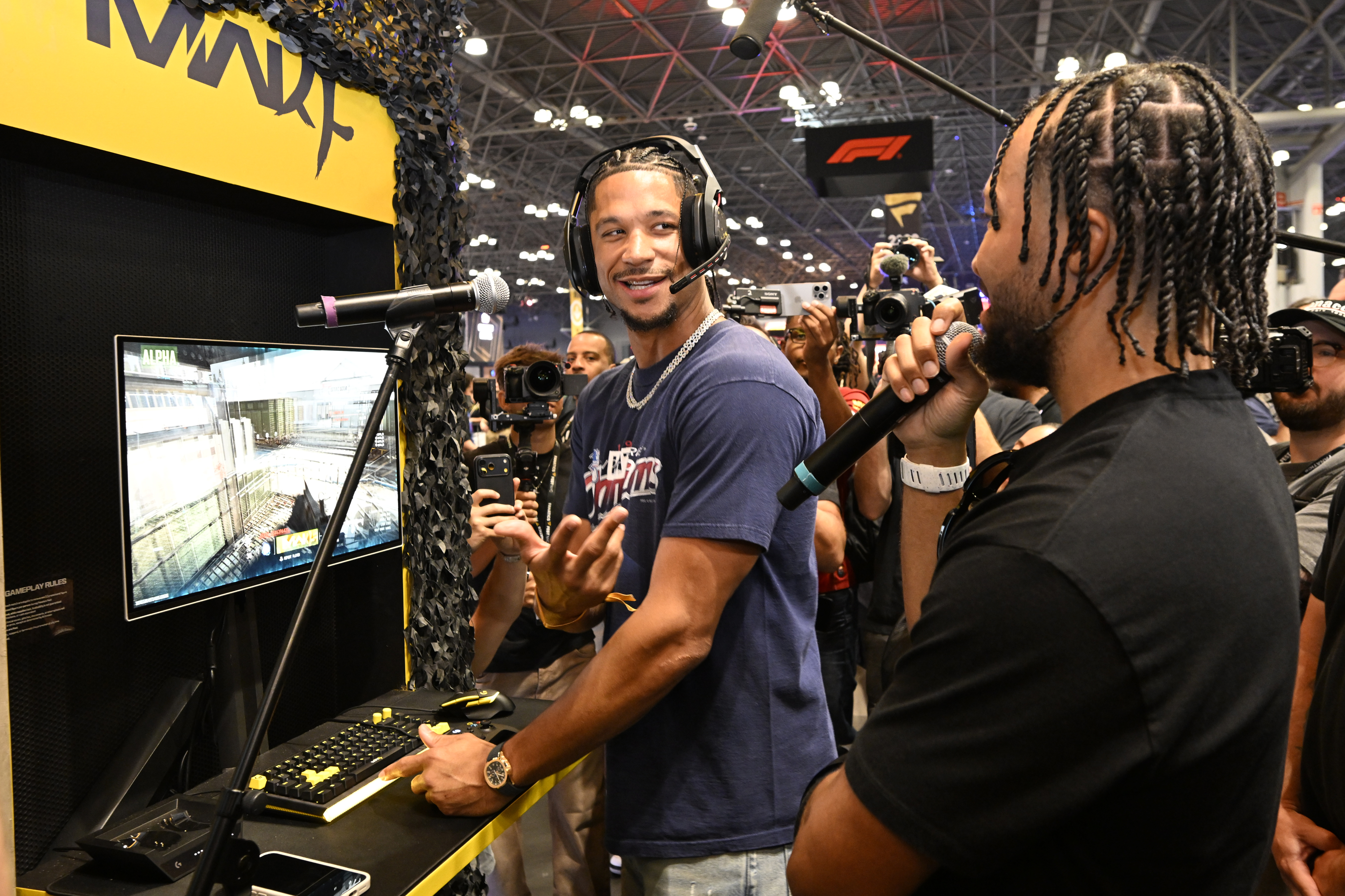 Two men, one wearing a headset, playing a video game on a large screen at a gaming event.